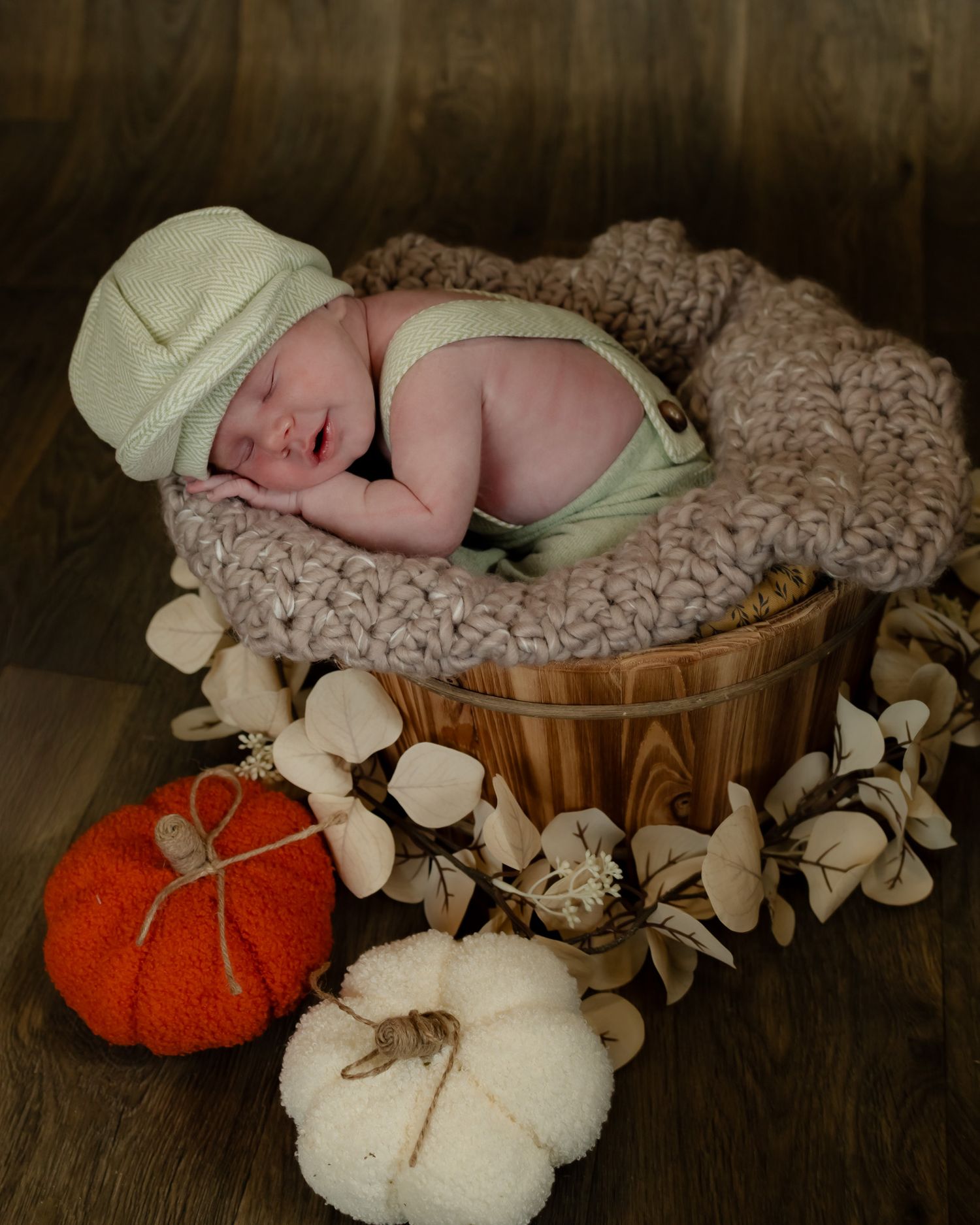A sleeping newborn baby rests in wooden buckets with soft knitted blankets and decorative white and orange pumpkins.