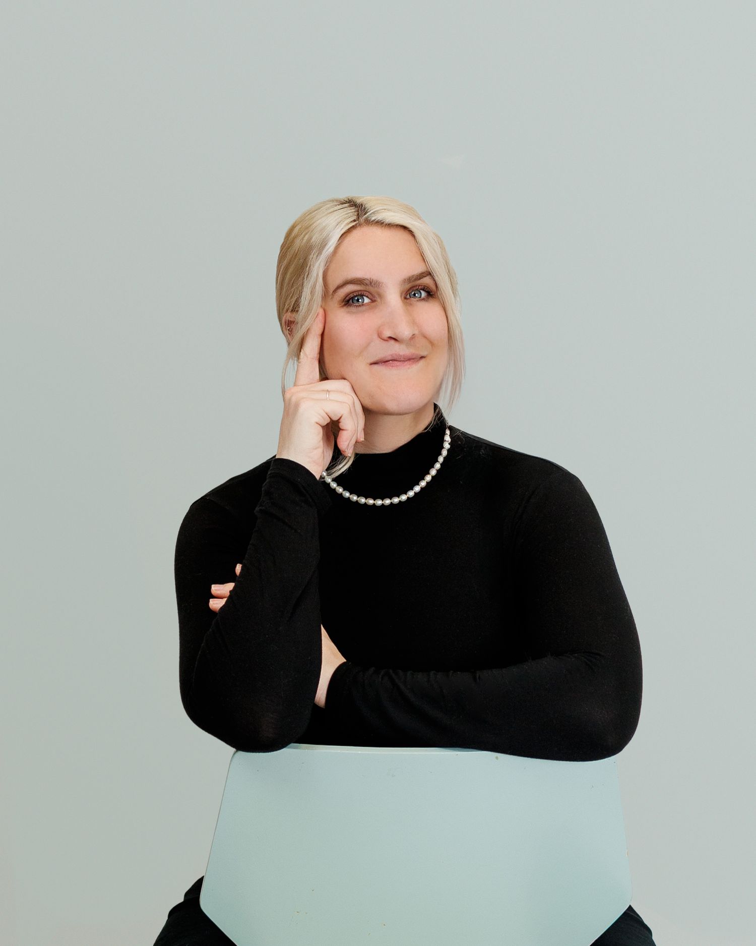 A figure in a black turtleneck sweater and pearl necklace poses against a light blue background in a series of artistic shots.