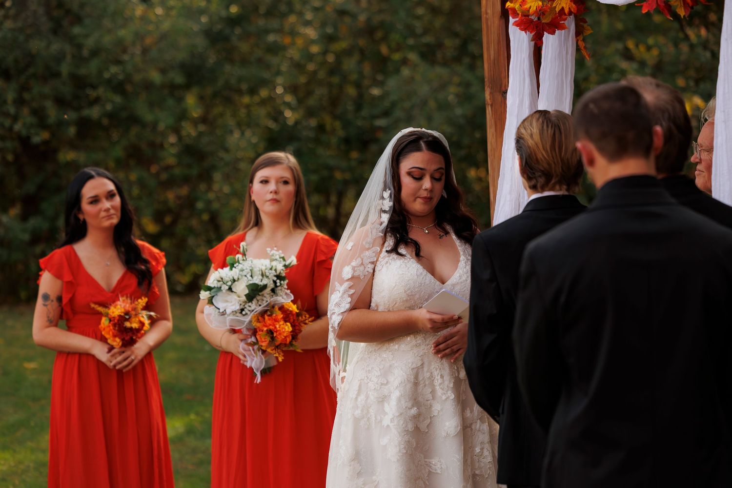 Wedding party members in bright red dresses and dark suits gather outdoors for formal photos during a fall ceremony.
