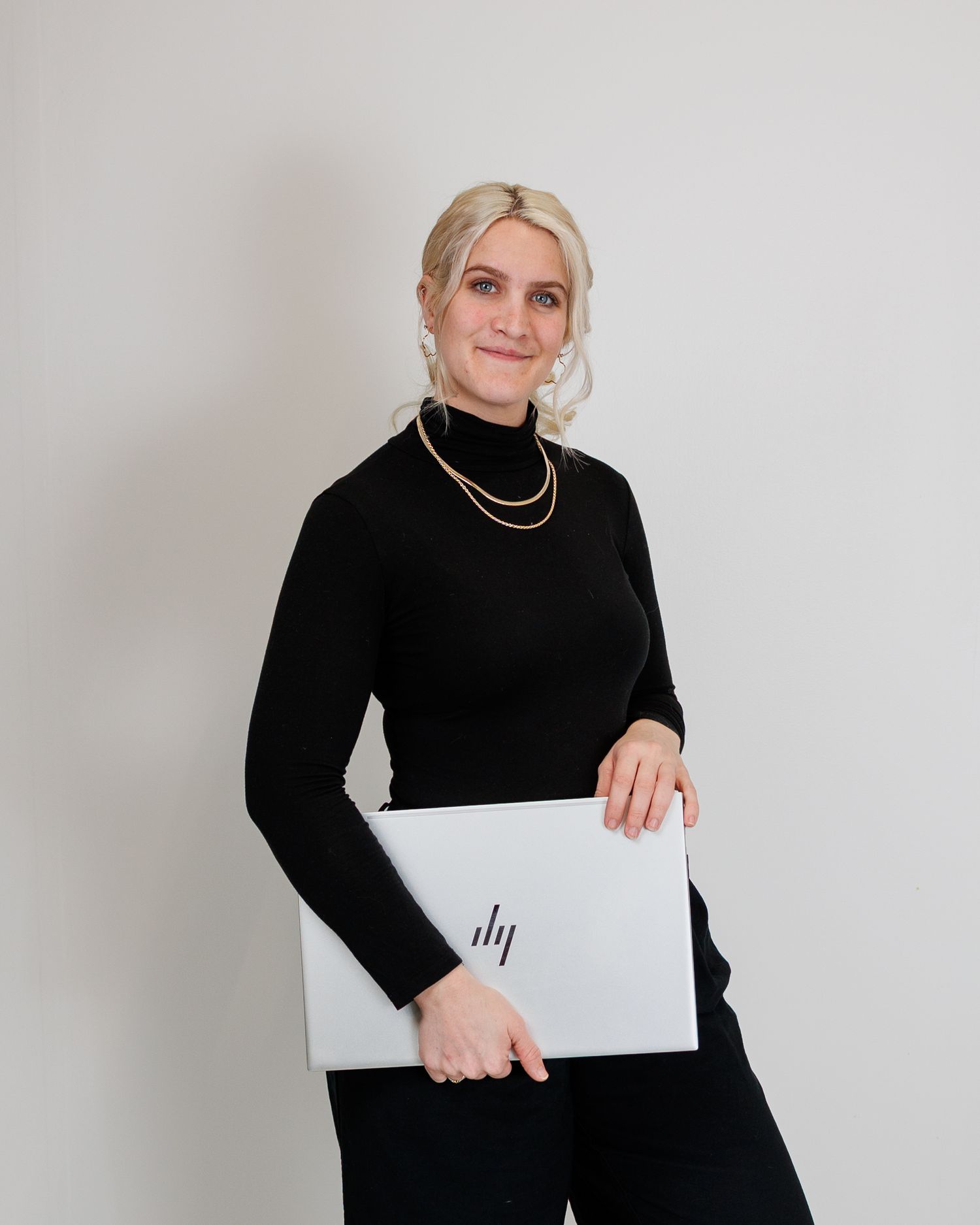 Professional in a black turtleneck sweater holding a silver laptop against a white wall.