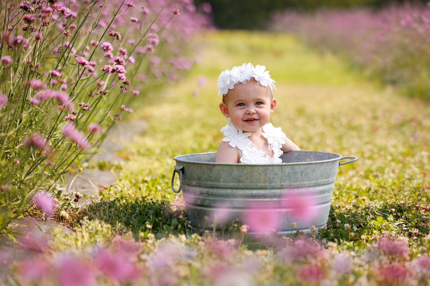 Smiling baby girl in white headband sitting in a galvanized tub surrounded by pink wildflowers outdoors.