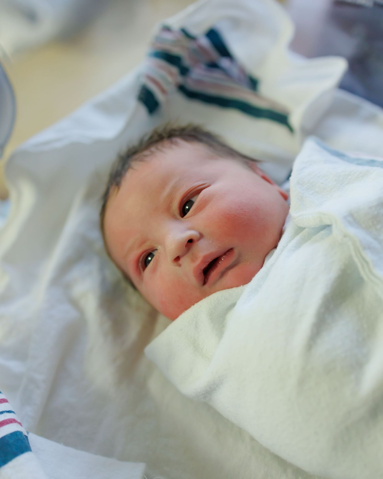Close-up of alert newborn baby in white clothing looking at camera.