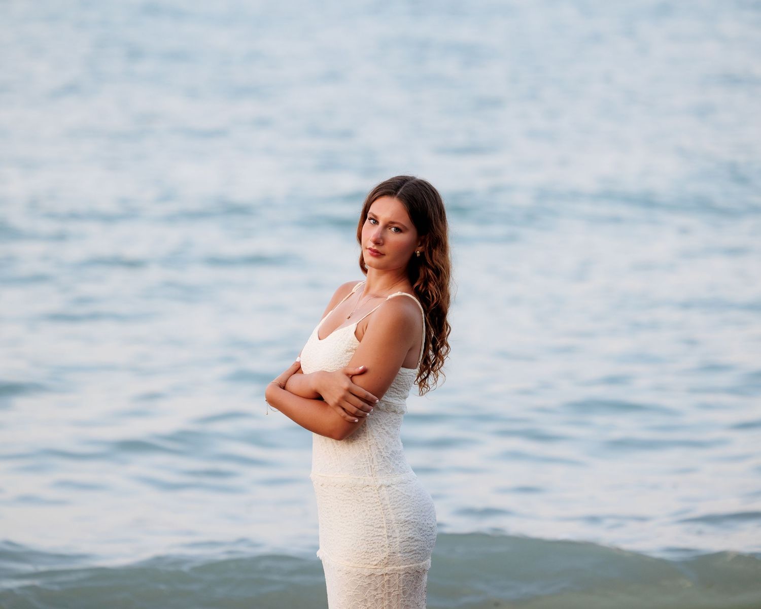 Woman in white lace dress posing gracefully by the ocean shore with arms crossed.