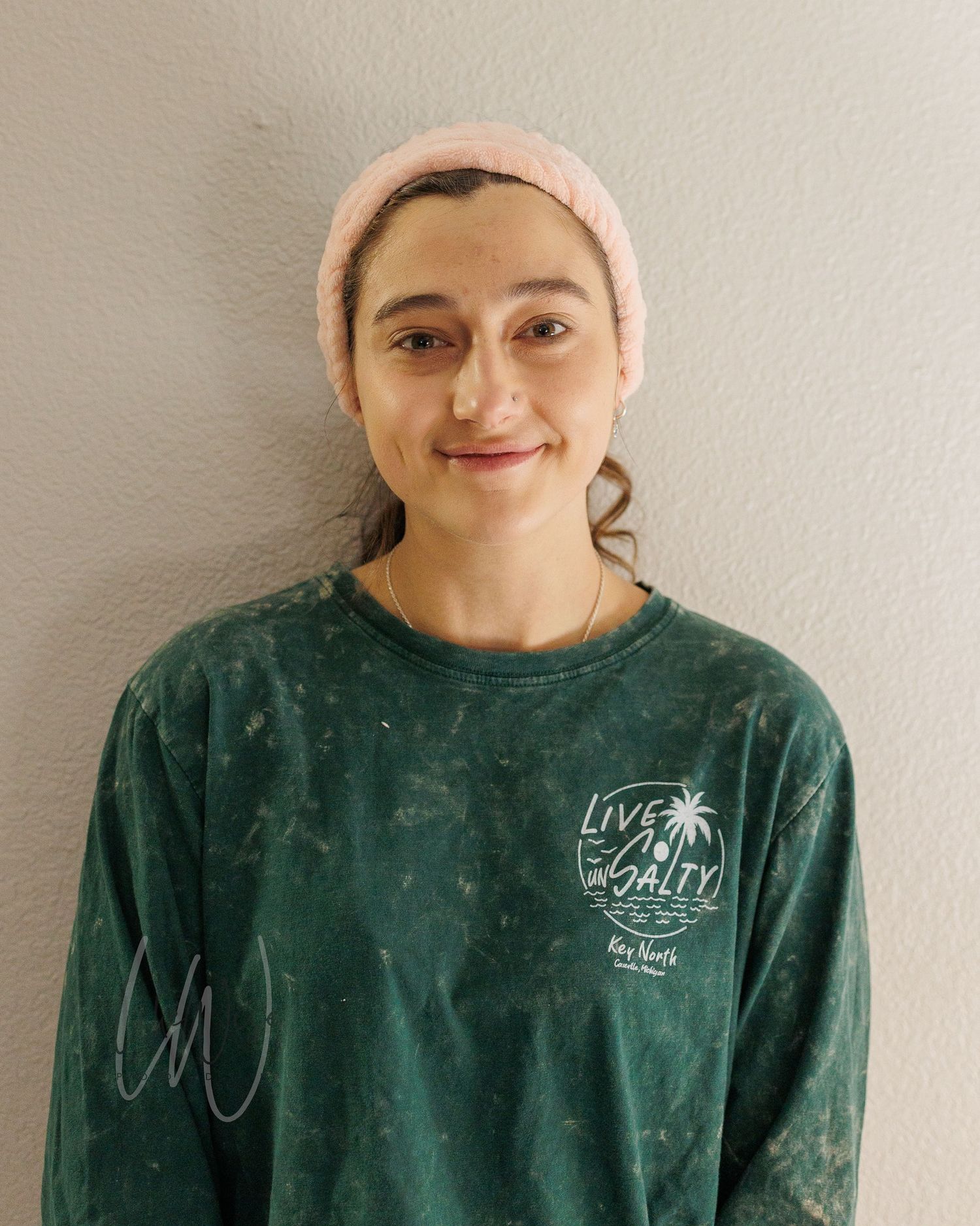 Someone wearing a pink beanie and forest green sweatshirt with a Palm tree logo stands against a white wall with a warm smile.