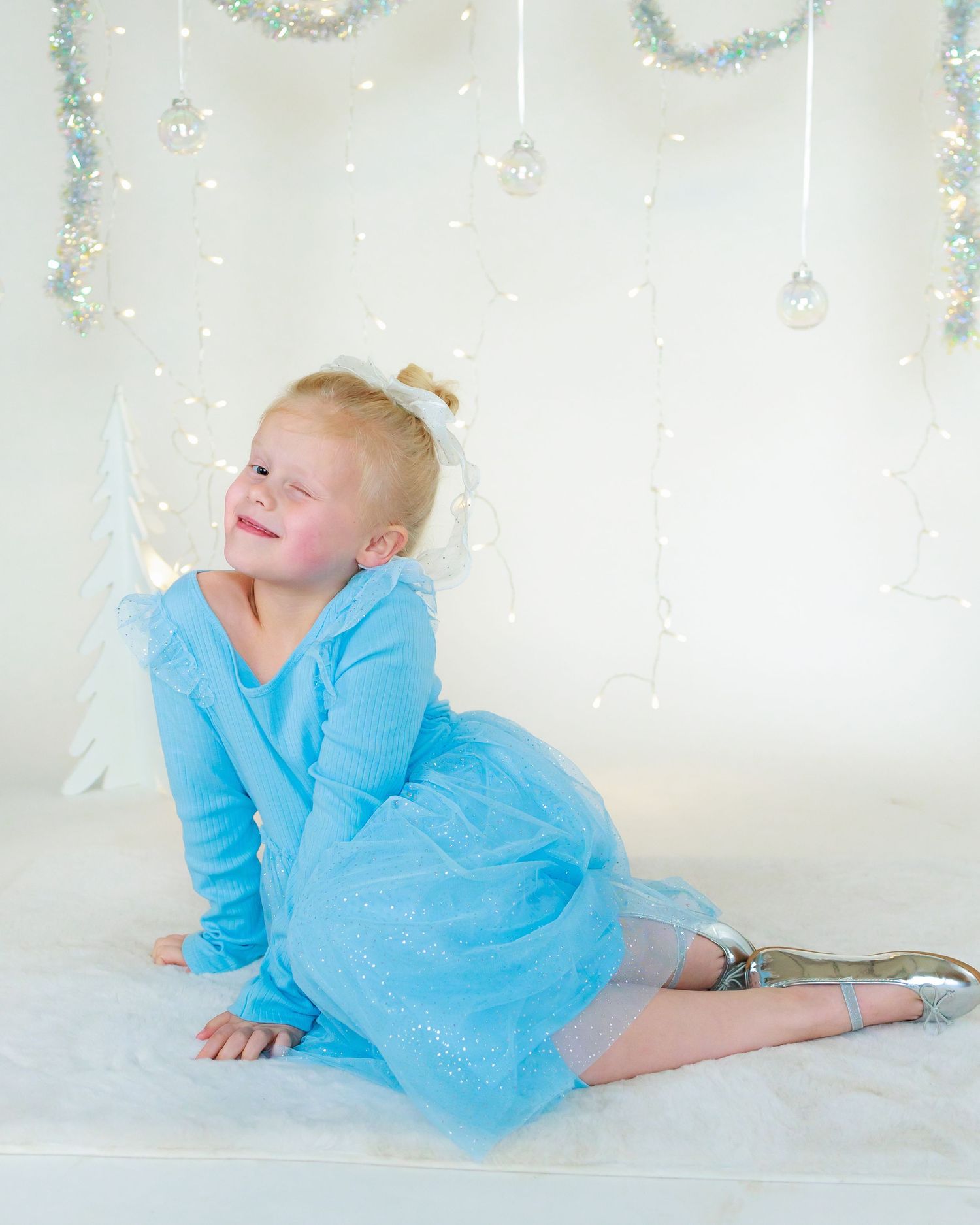 Child in light blue dress poses against white backdrop with hanging decorative elements.