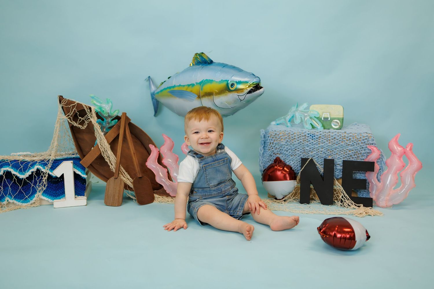Baby boy sits smiling amid fishing-themed first birthday decor with fish balloon, nets, and ONE sign on blue backdrop.