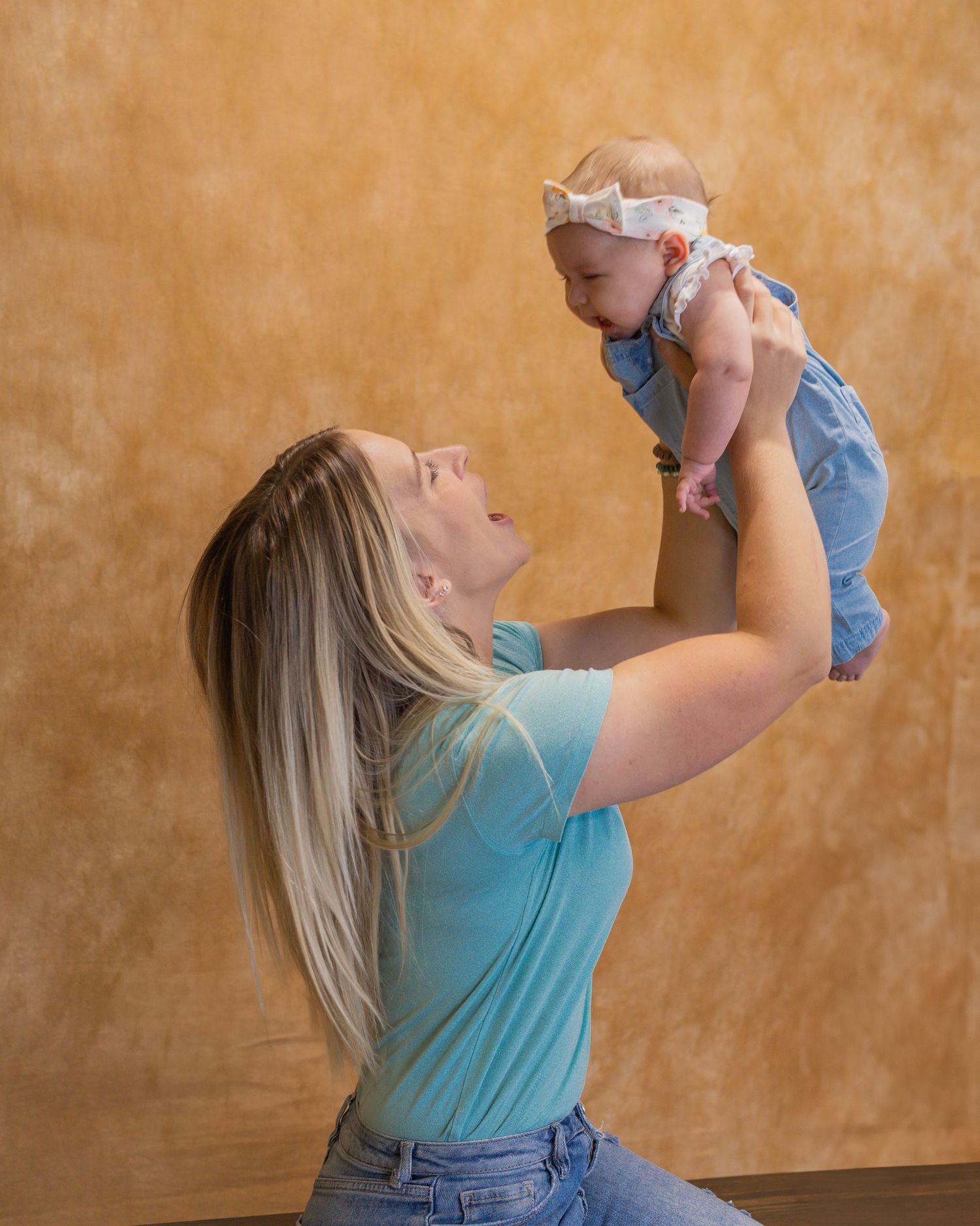 Mother in blue shirt lifting baby wearing denim overalls against warm orange background.