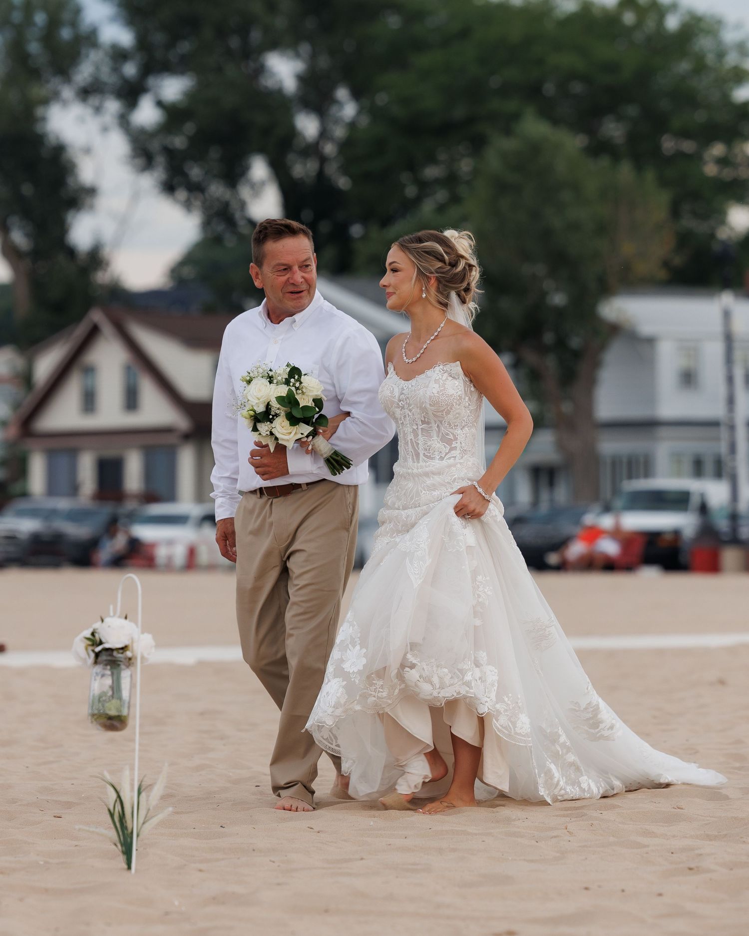 A couple on a beach during a wedding photoshoot, with the bride wearing a lace gown and groom in casual attire.