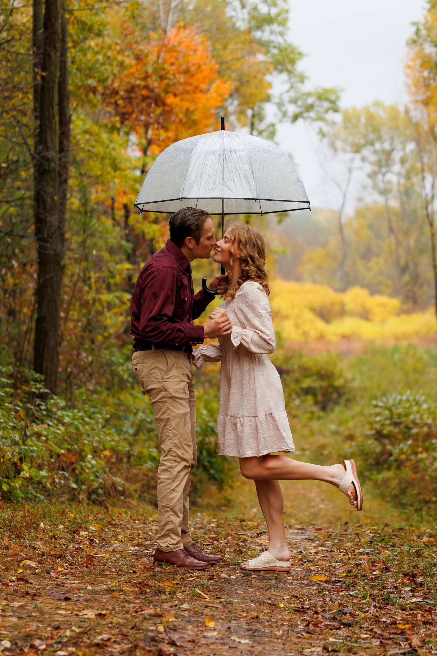 A romantic autumn scene shows a couple sharing a tender moment under an umbrella on a leaf-covered forest path.