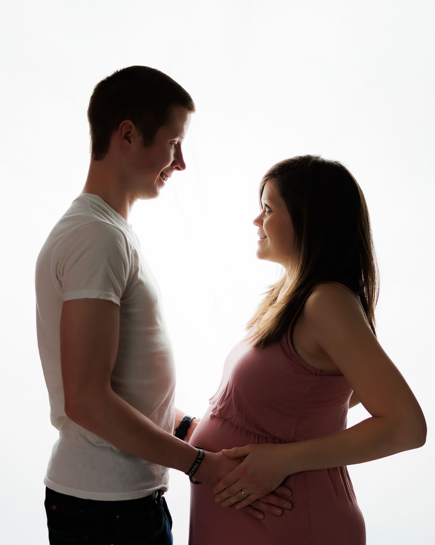 Silhouetted couple in white t-shirt and pink dress share an intimate moment during maternity photoshoot against light background.