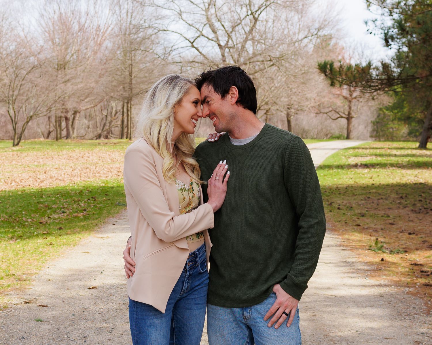 Two people share an intimate moment while standing together on a path surrounded by winter trees in a park.