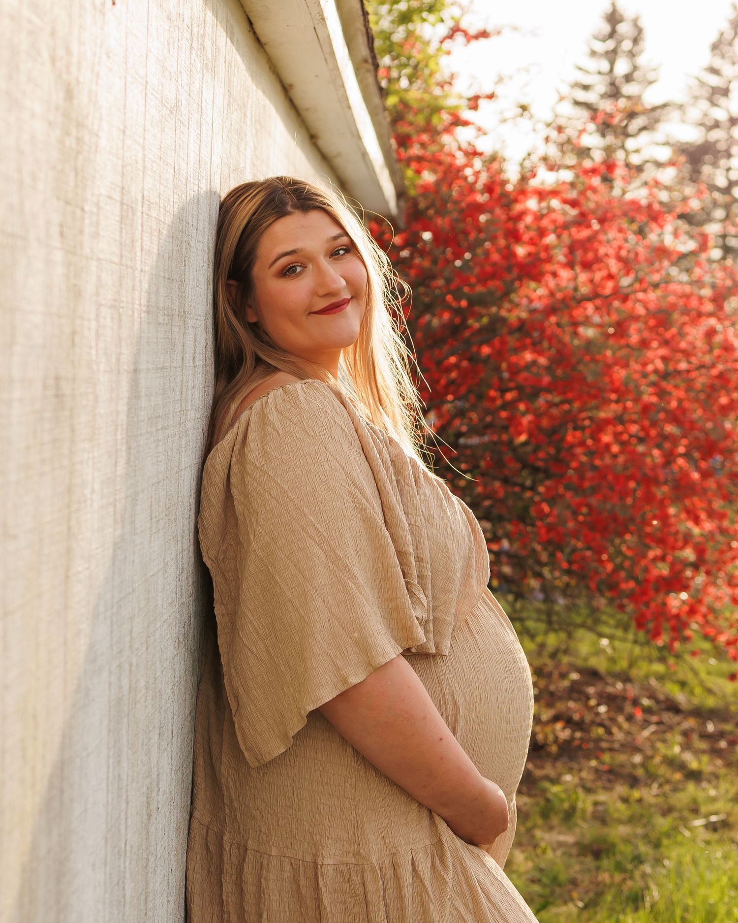 Someone in a beige dress poses against a white wall with vibrant red autumn foliage in the background on a sunny day.
