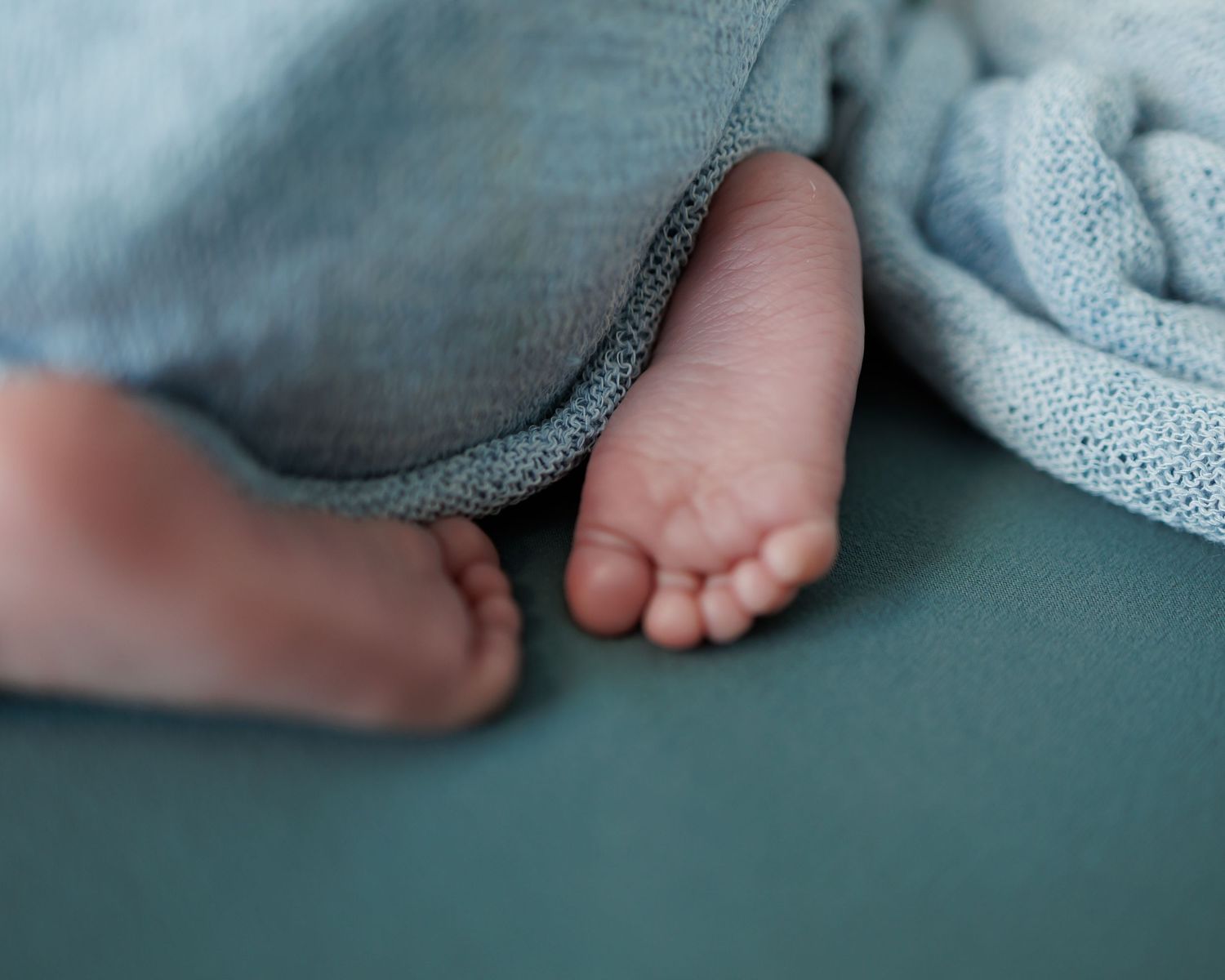 Tiny baby feet peek out from under a soft blue blanket on a teal surface.