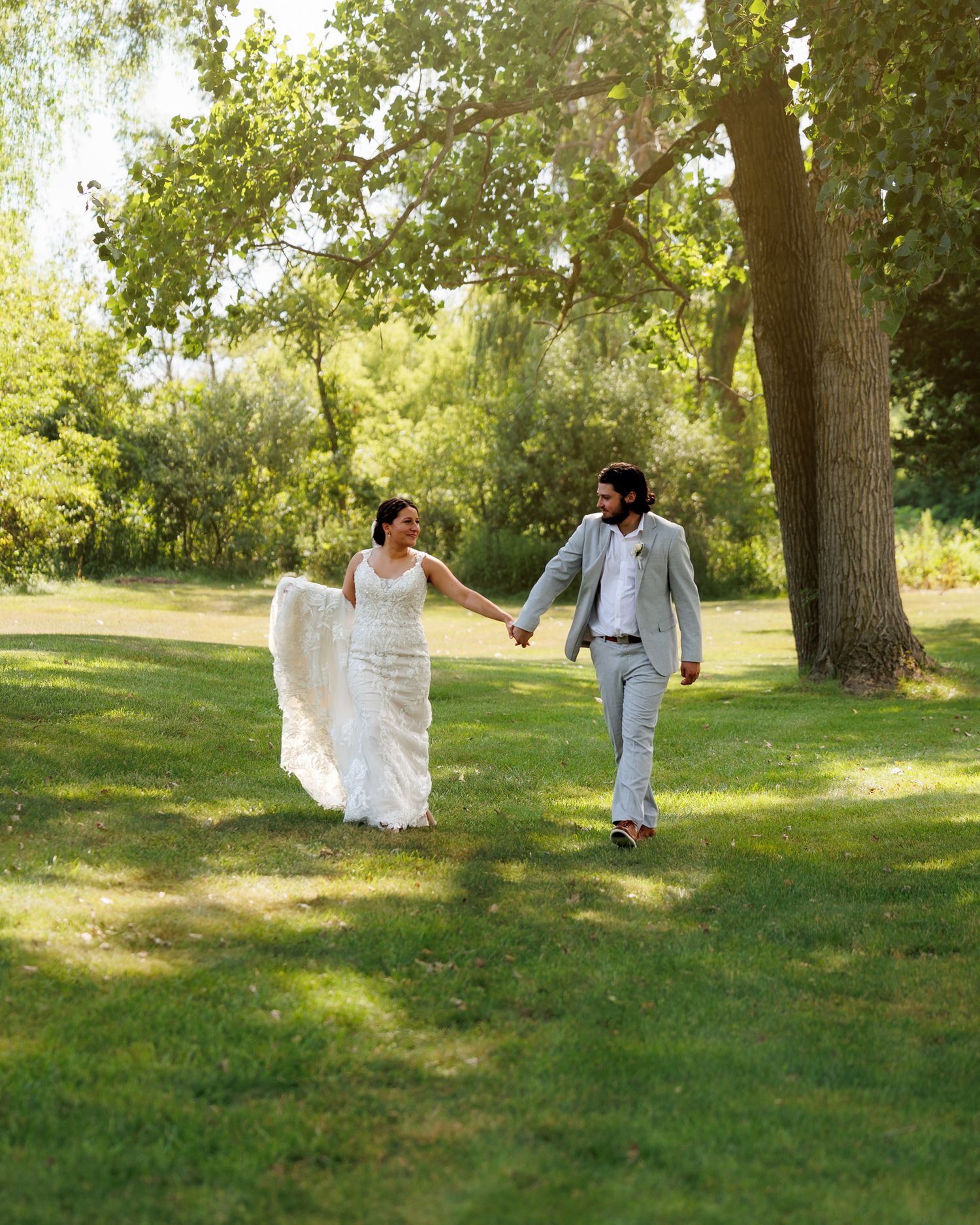 Bride and groom holding hands walking through lush green park on sunny wedding day.