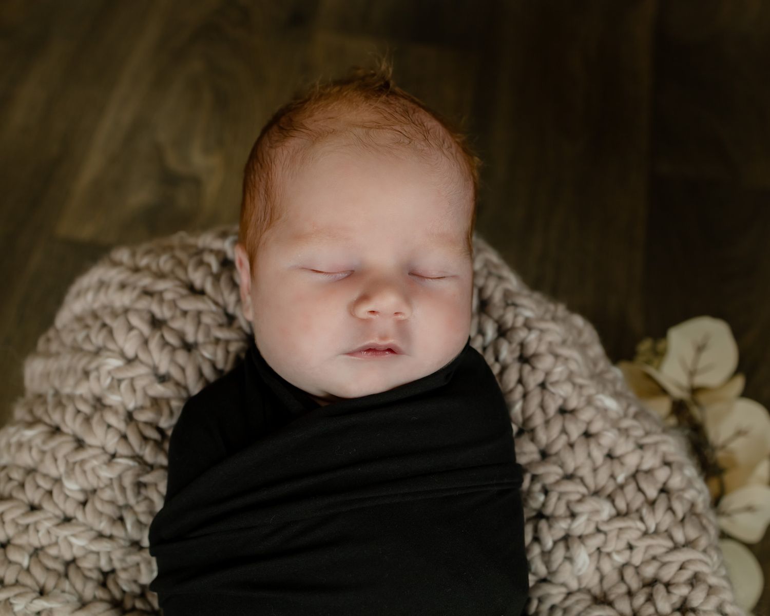 Sleeping newborn wrapped in black swaddle on textured beige blanket.
