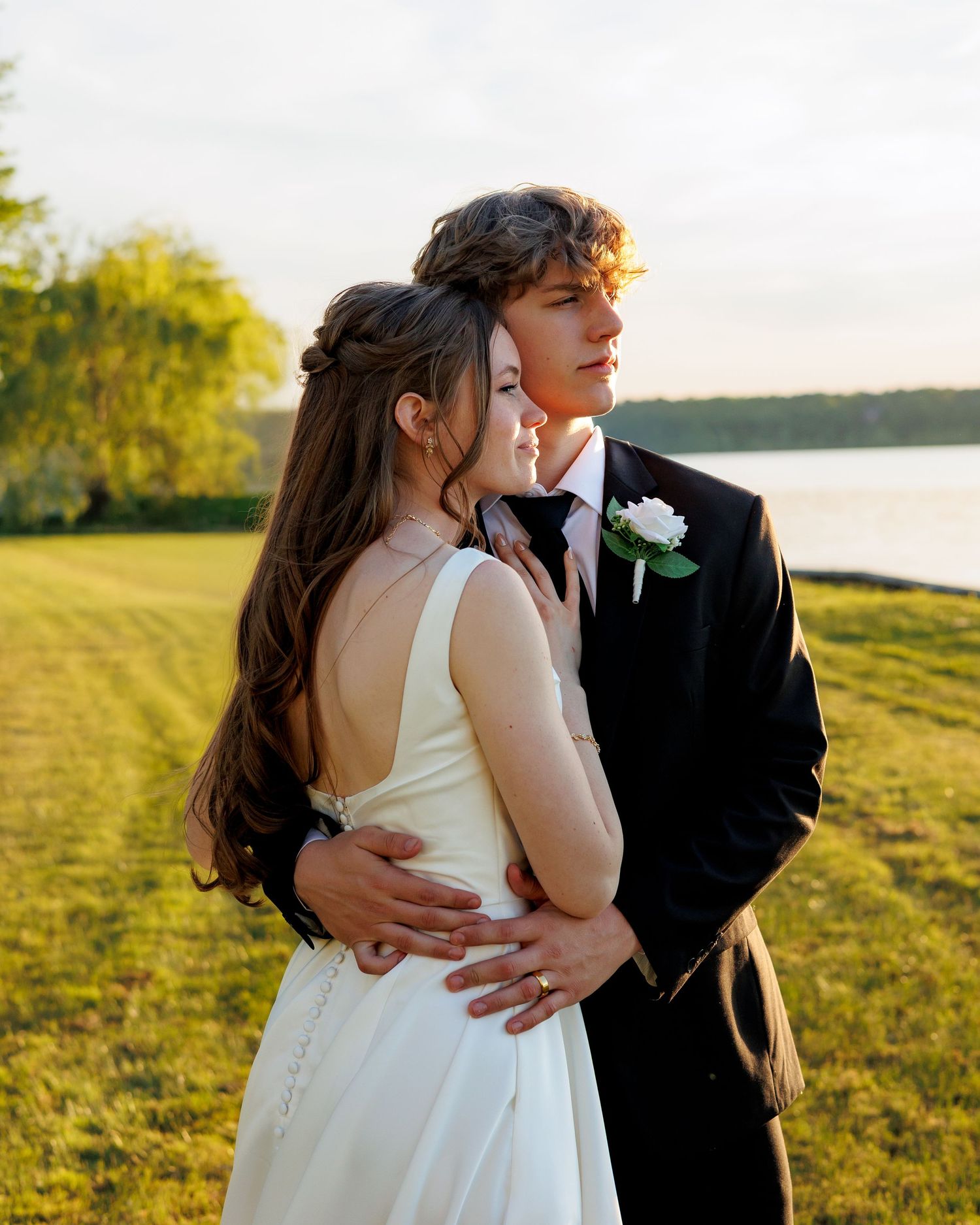 Young couple in formal wear embracing outdoors at golden hour near a lake, woman in white dress, man in black suit.