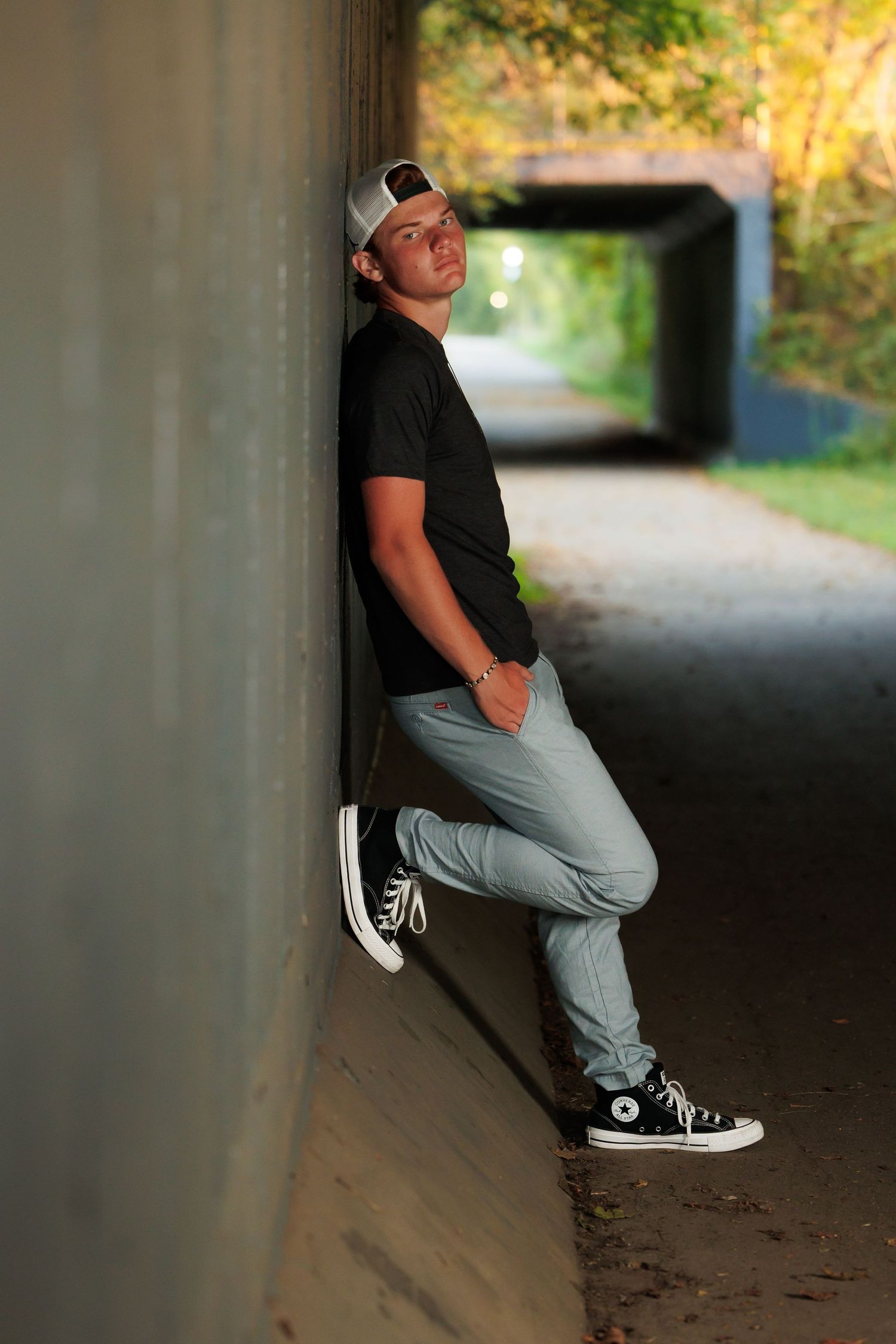 A casual portrait of someone leaning against a tunnel wall wearing a baseball cap, black t-shirt and Converse sneakers.