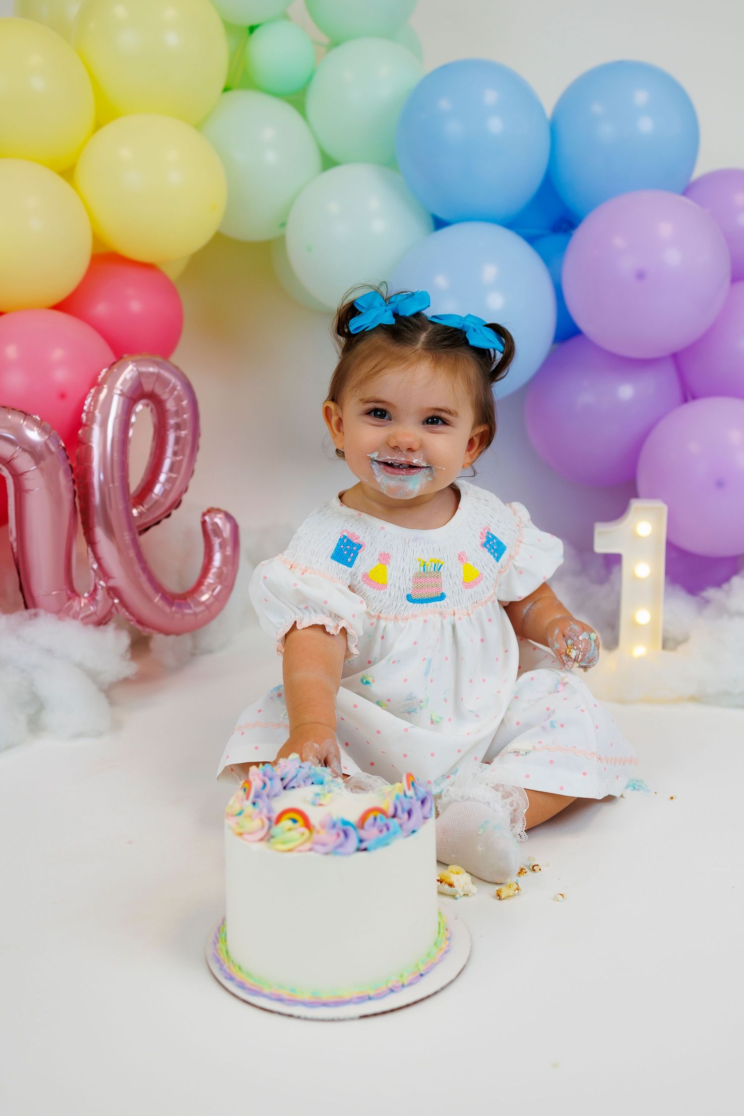 First birthday cake smash photo session with pastel rainbow balloon arch and white frosted cake with number one candle.