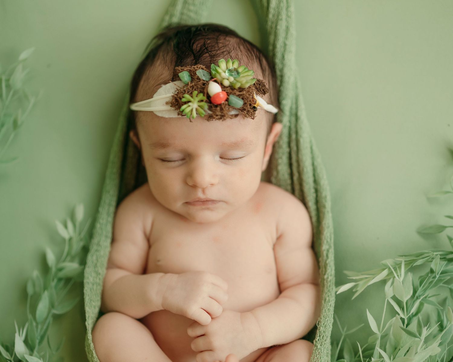 A sleeping newborn baby wearing a delicate floral headband lies on soft green fabric in a professional photography setting.