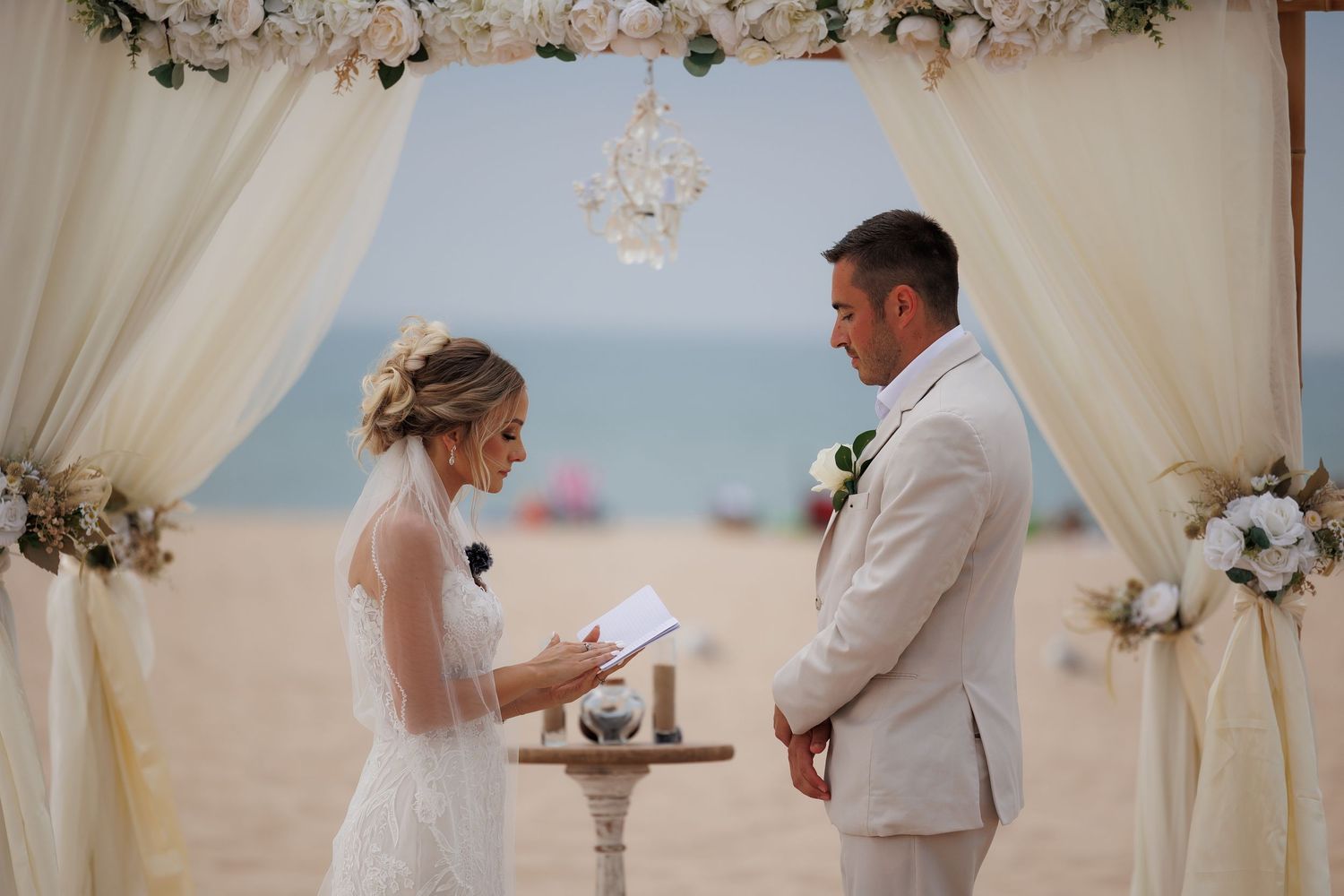 Bride and groom exchange rings during a romantic beach wedding ceremony under white floral decorations.