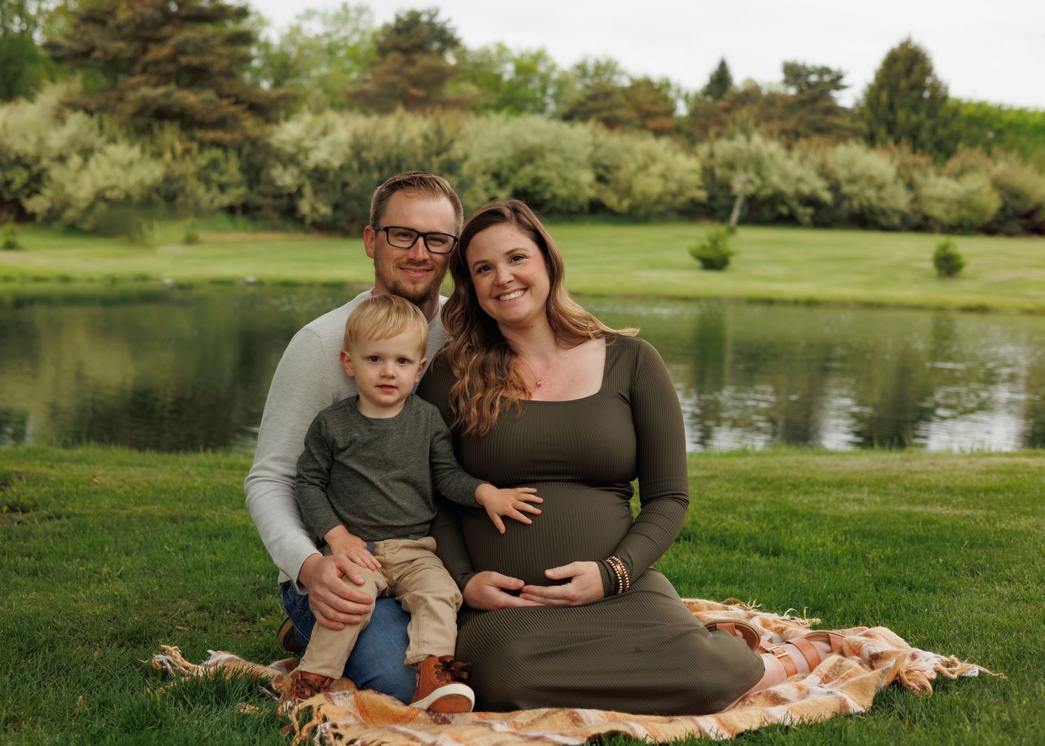A family sits on a picnic blanket by a tranquil pond surrounded by green trees and grass.
