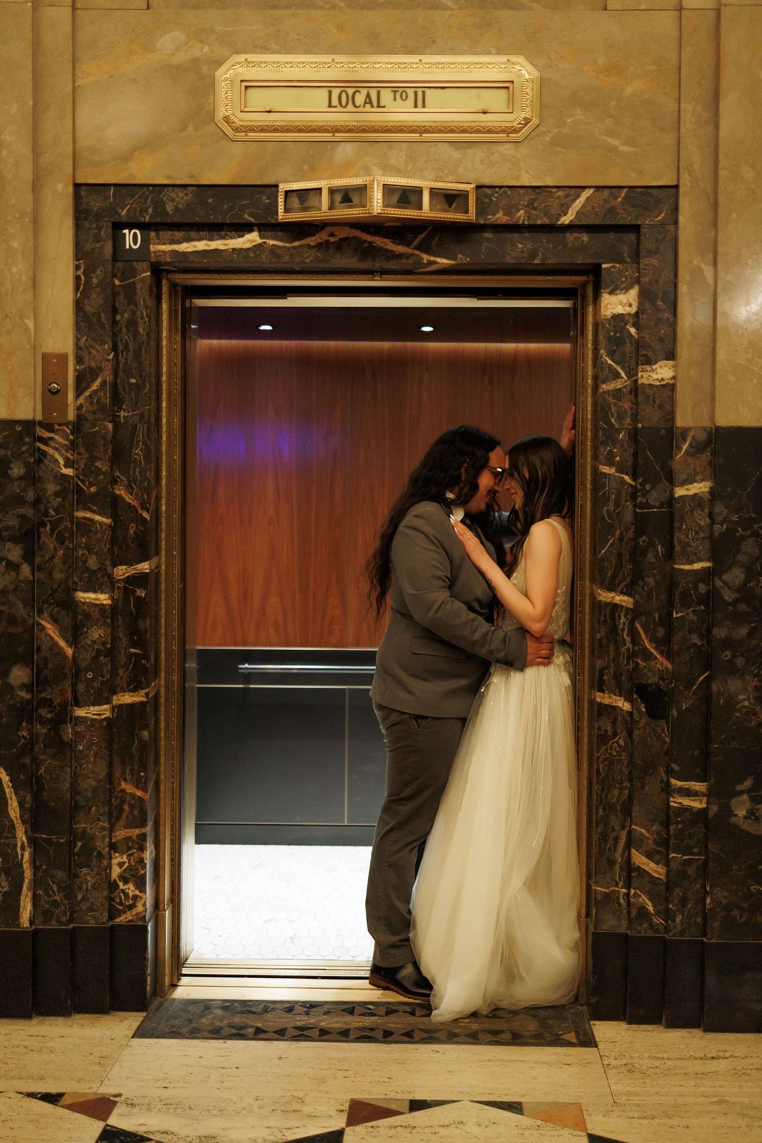 A romantic wedding couple shares a tender kiss in front of an elevator with marble walls in an art deco building.