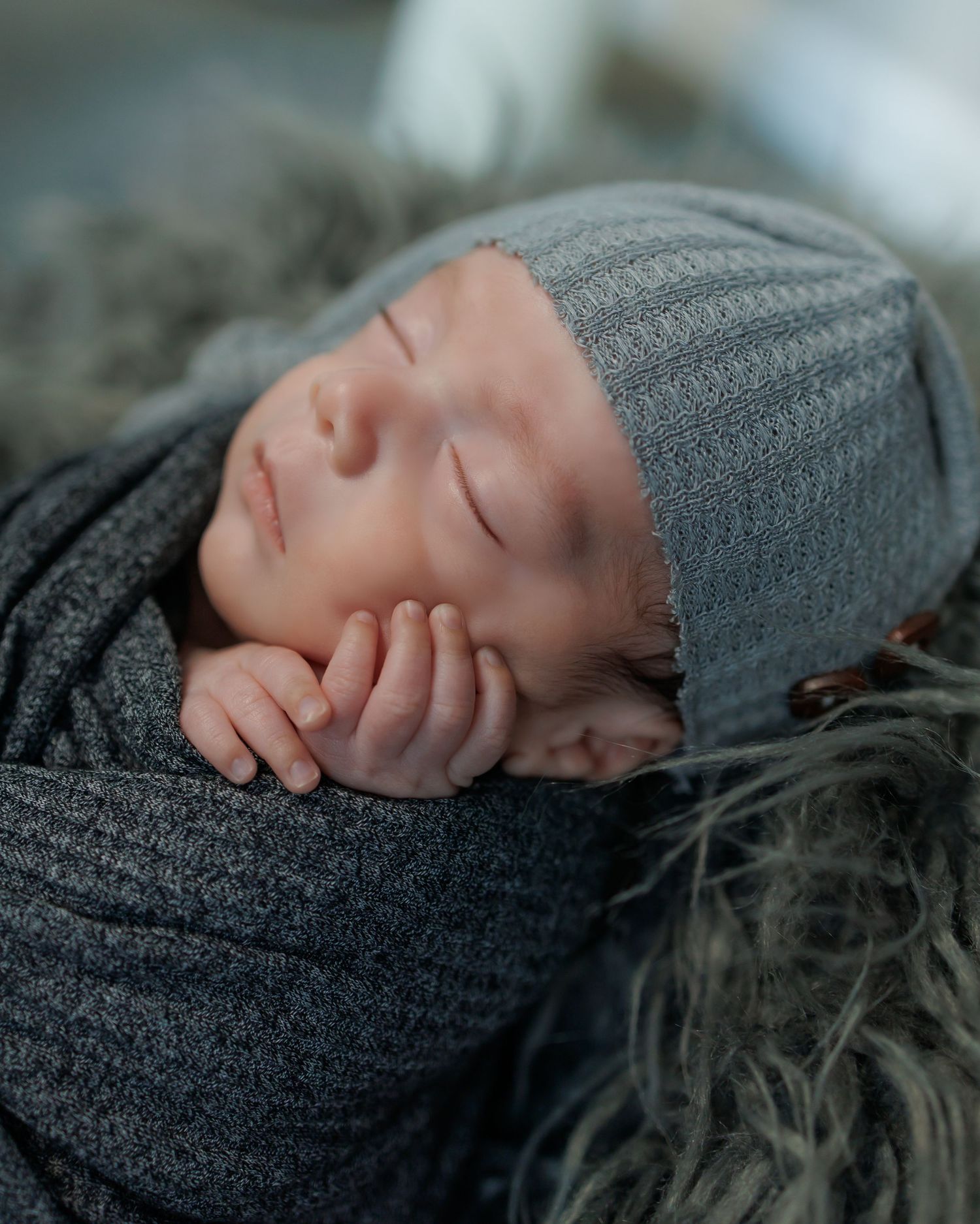 Sleeping baby wrapped in soft gray knit blanket and wearing matching hat rests peacefully on furry surface.