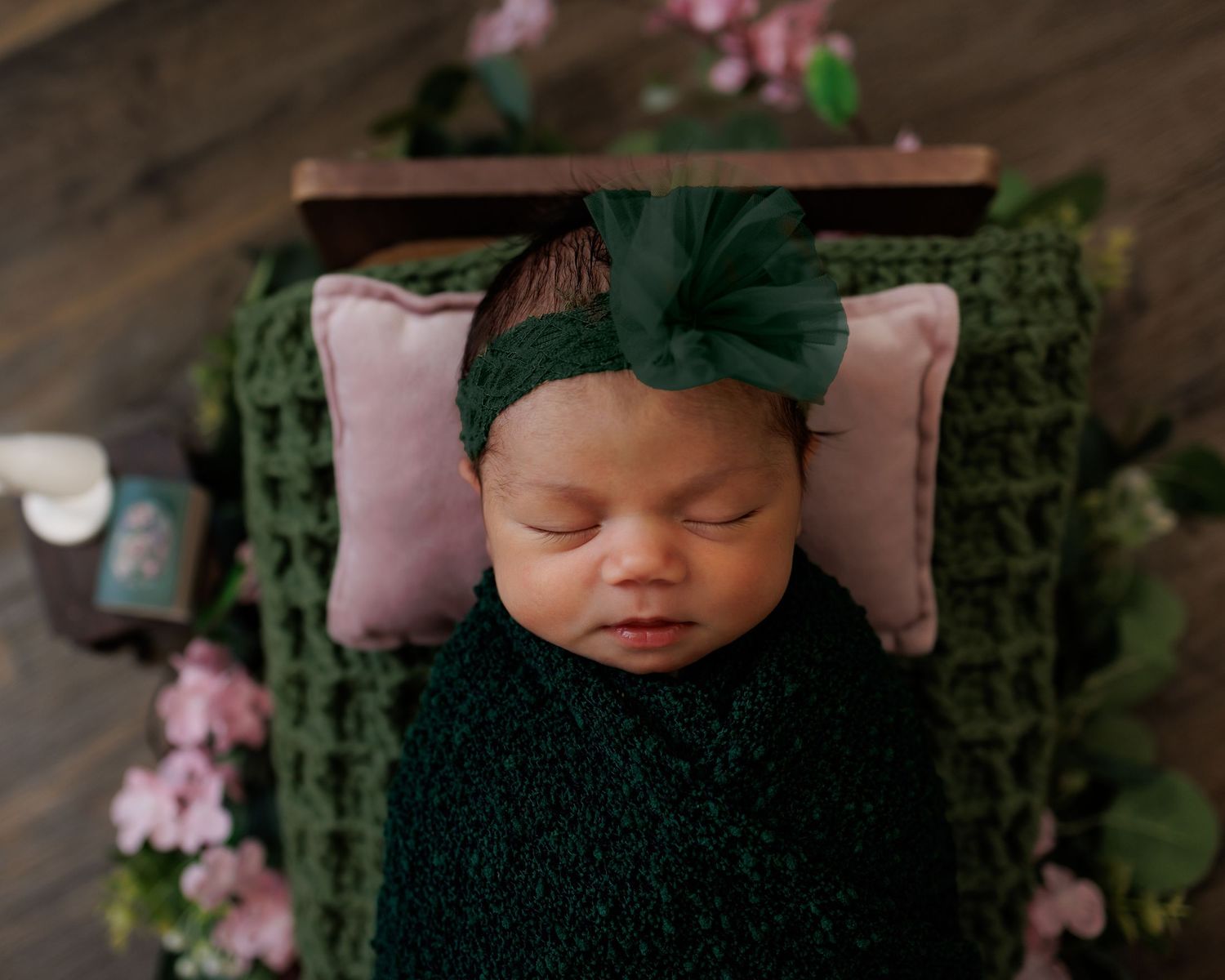 Newborn baby swaddled in dark green wrap with floral headband, sleeping peacefully on pink pillow with flower decorations.