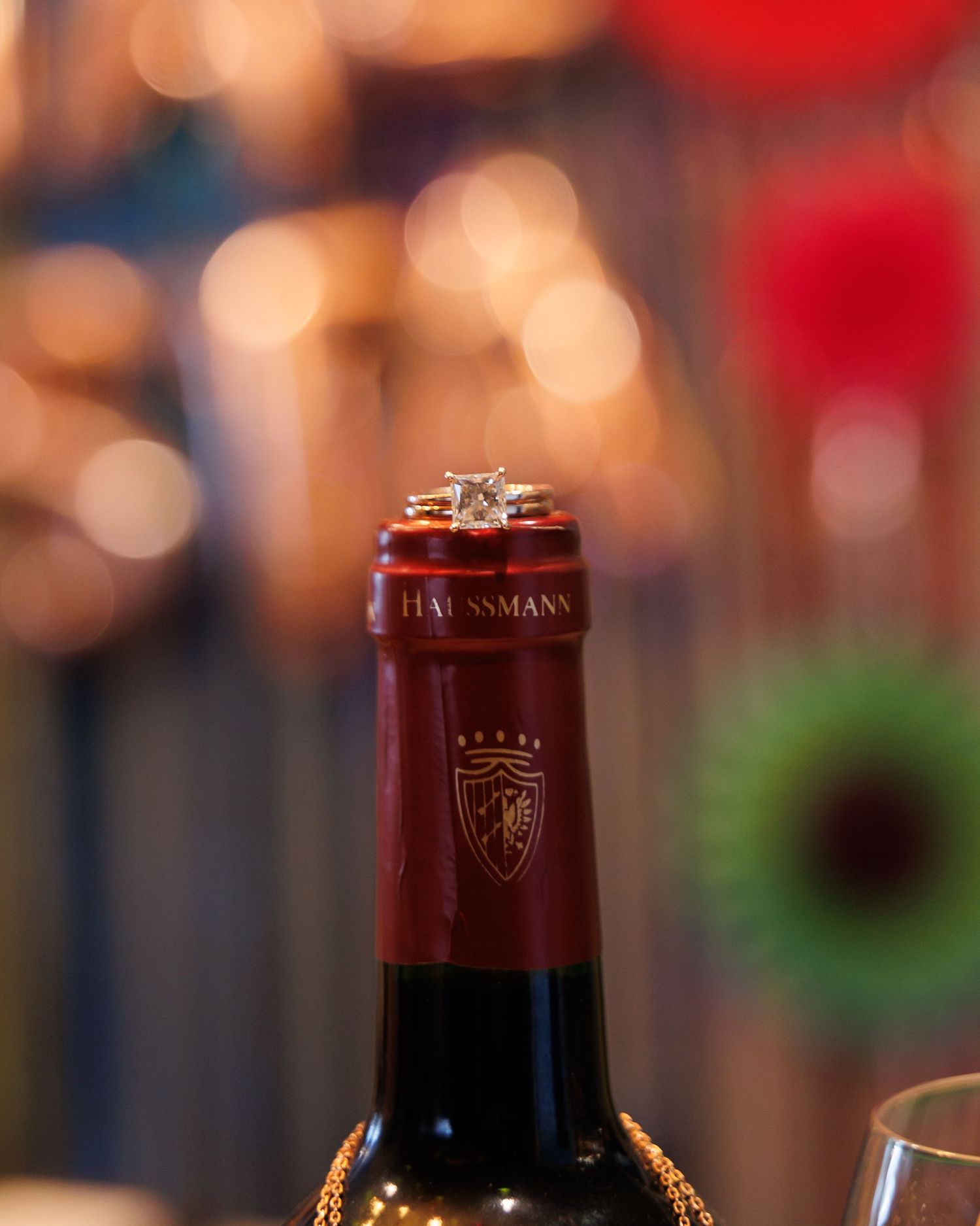 Wedding rings perched on top of a wine bottle with a blurred festive background of bokeh lights.