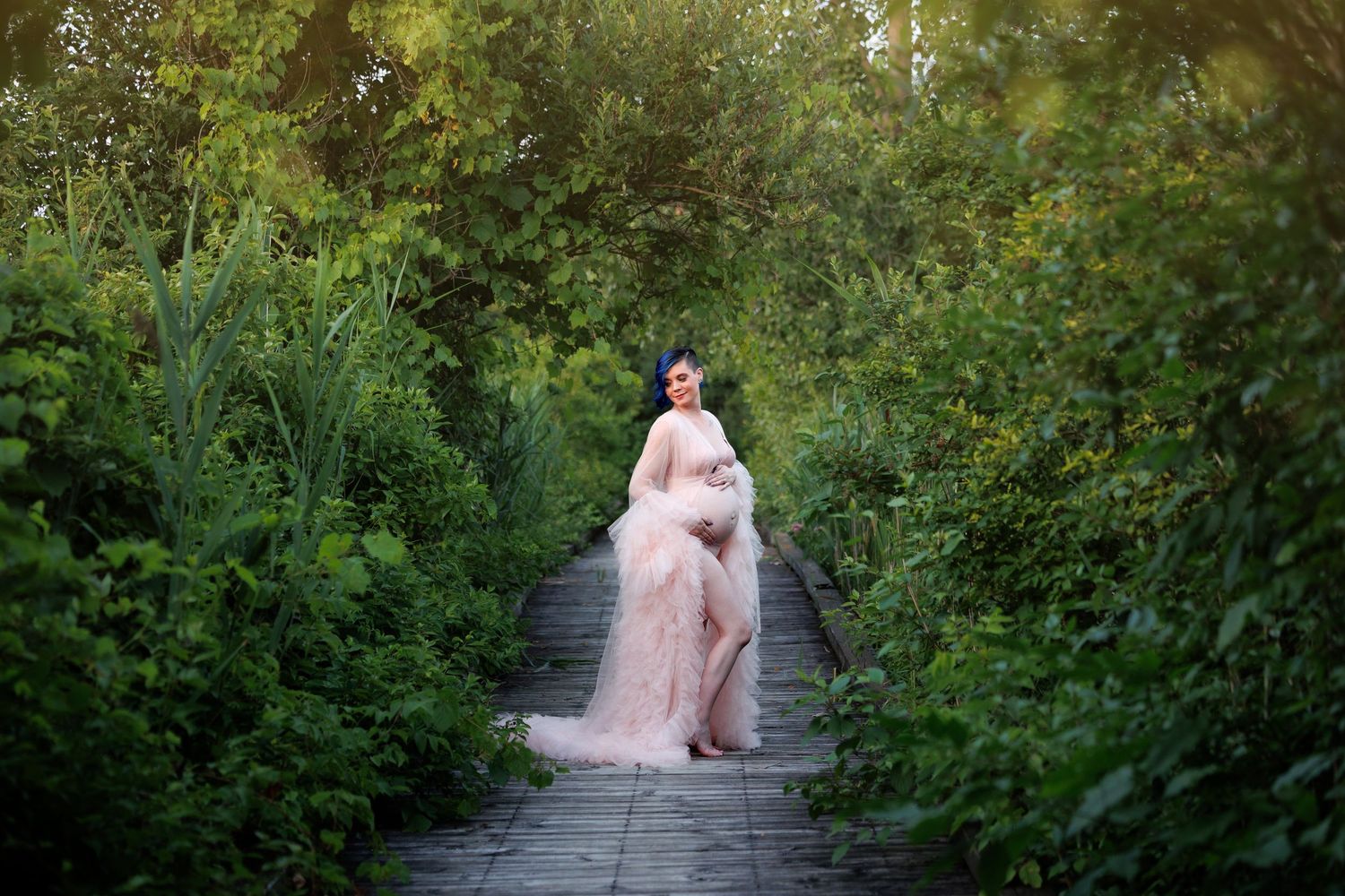 Pregnant woman in pink tulle gown poses on garden path surrounded by lush green foliage during golden hour.