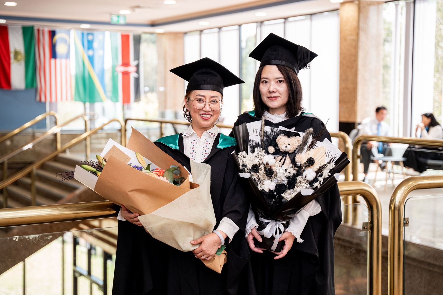 Two graduates in black caps and gowns holding bouquets stand together at a graduation ceremony with flags in background.