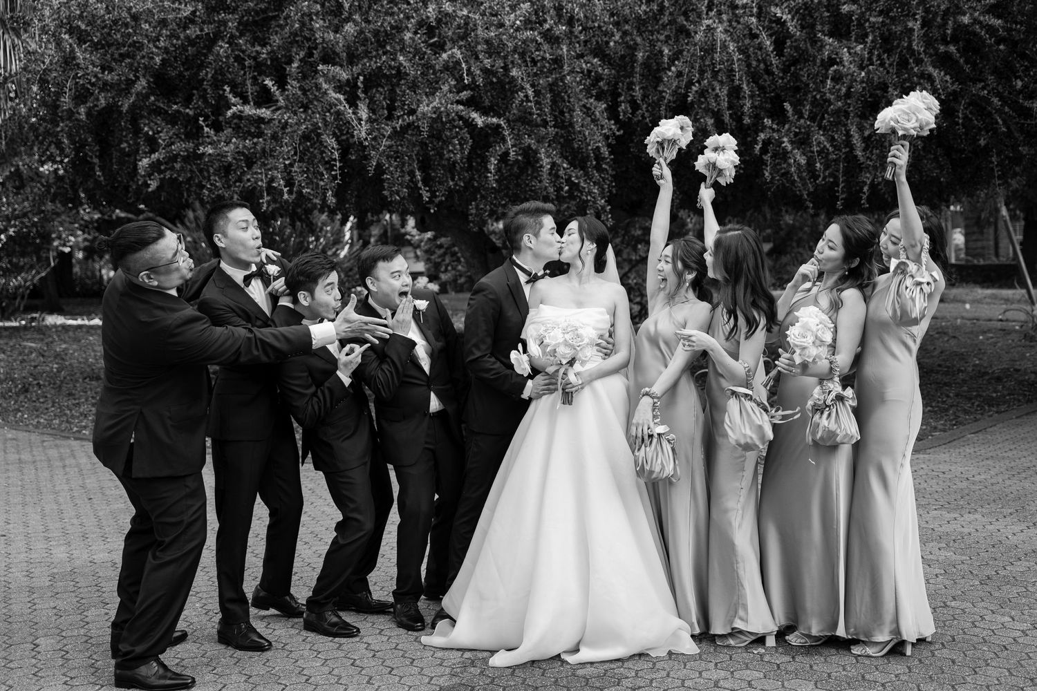 Black and white photo of wedding party posing outdoors with groomsmen leaning to one side and bridesmaids tossing bouquets.