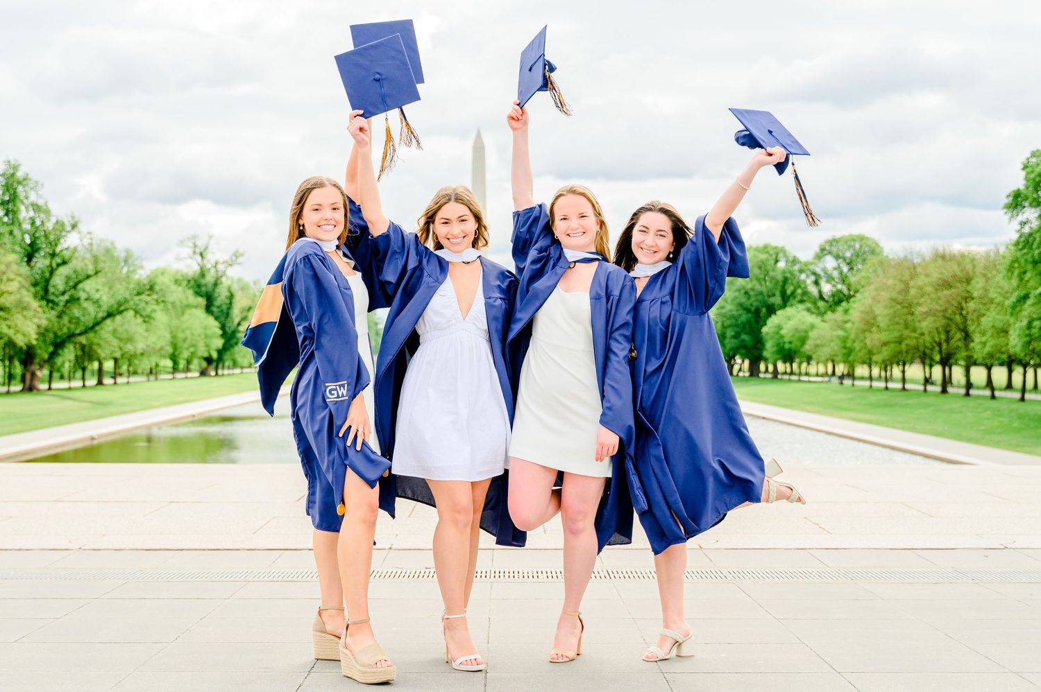 Four graduates in blue gowns pose together on campus grounds during celebration.
