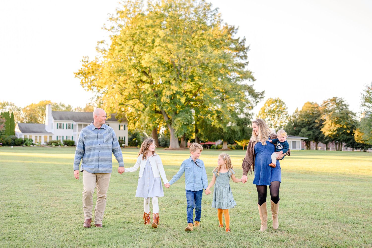 Family of six walking hand-in-hand across a sunlit green lawn with golden autumn trees and a historic estate in the background.