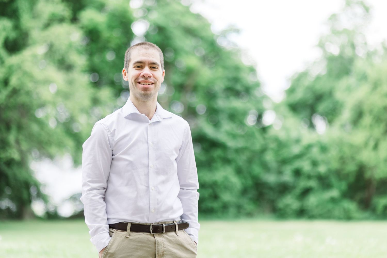 Professional headshot of someone in a light button-down shirt and khaki pants standing outdoors with green foliage.