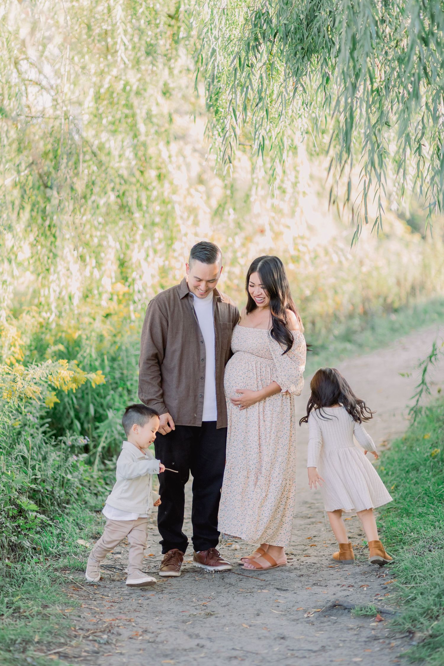A family walks together on a sunlit dirt path under willow trees in a natural outdoor setting.