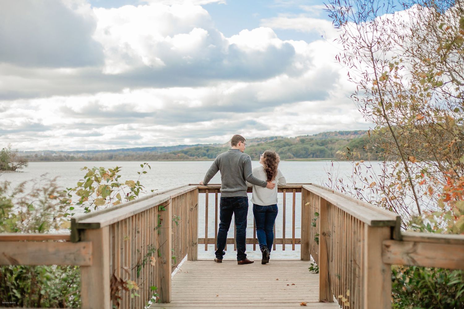 A couple stands together on a wooden boardwalk overlooking a scenic lake with cloudy skies and autumn trees.
