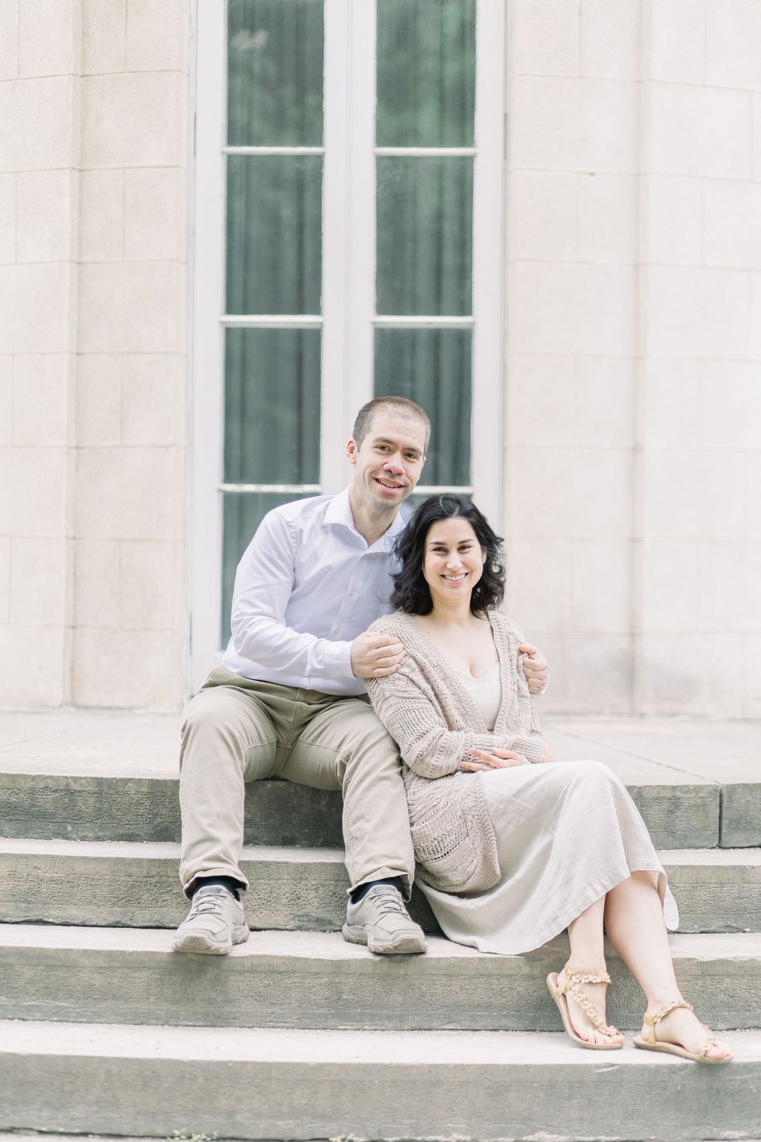A couple sits together on concrete steps outside a building with tall windows, dressed in light neutral colors.