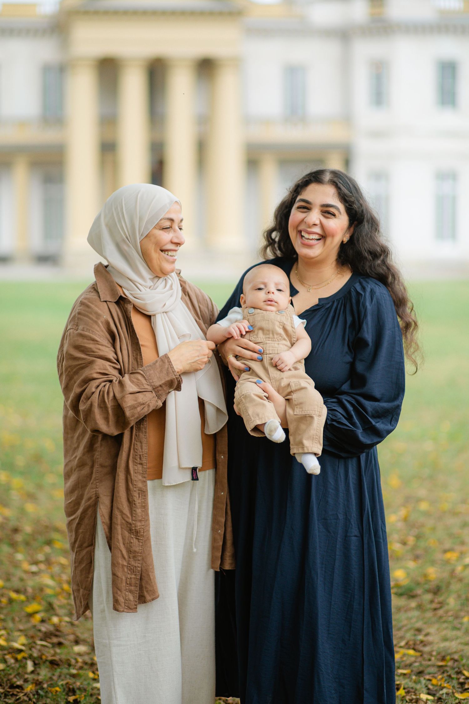 Three generations gather outdoors on an autumn day with fallen leaves on the ground near a stately building.