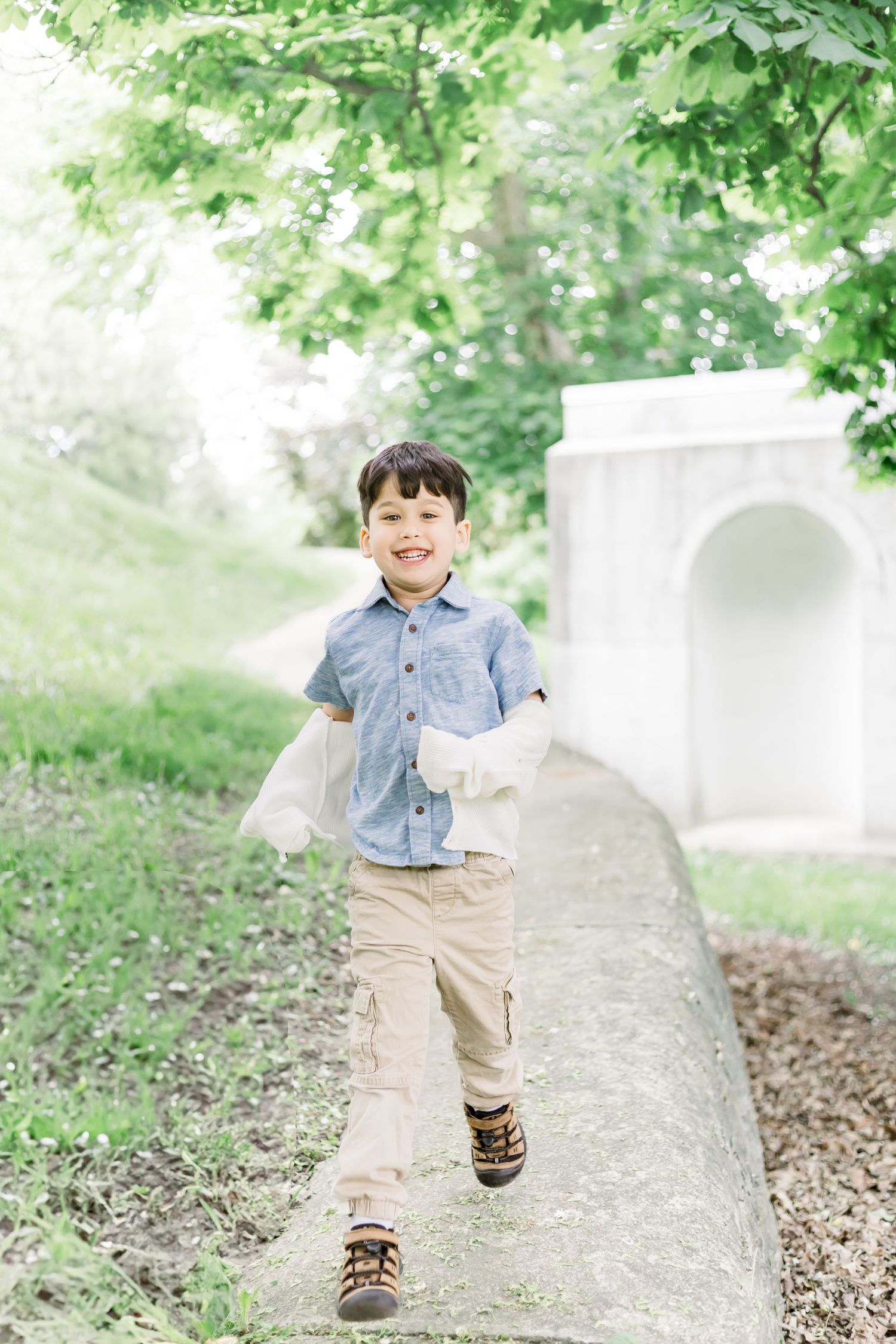A child in denim shirt and khaki pants plays and walks along a stone wall in a lush green garden setting.