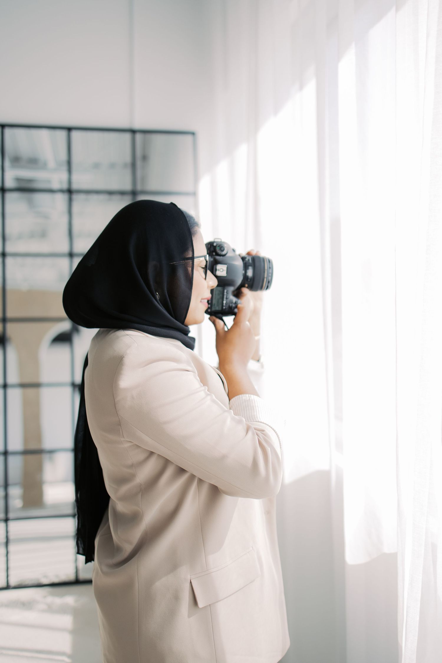 A photographer wearing a white blazer and black hijab takes photos in a bright, minimalist studio space.
