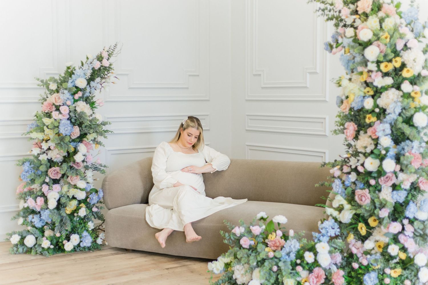 A bride in an elegant white gown poses beside a floral arch with pastel flowers and soft natural lighting.