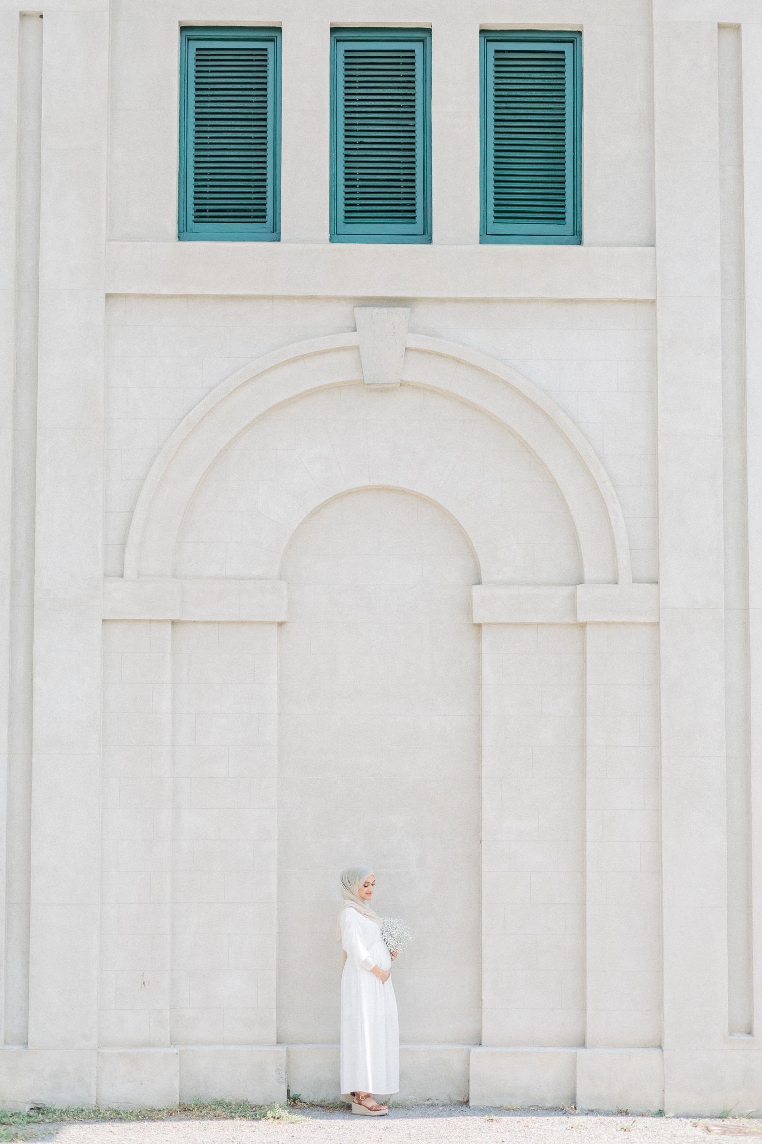 Small figure in white stands against a minimalist white building facade with turquoise shuttered windows.