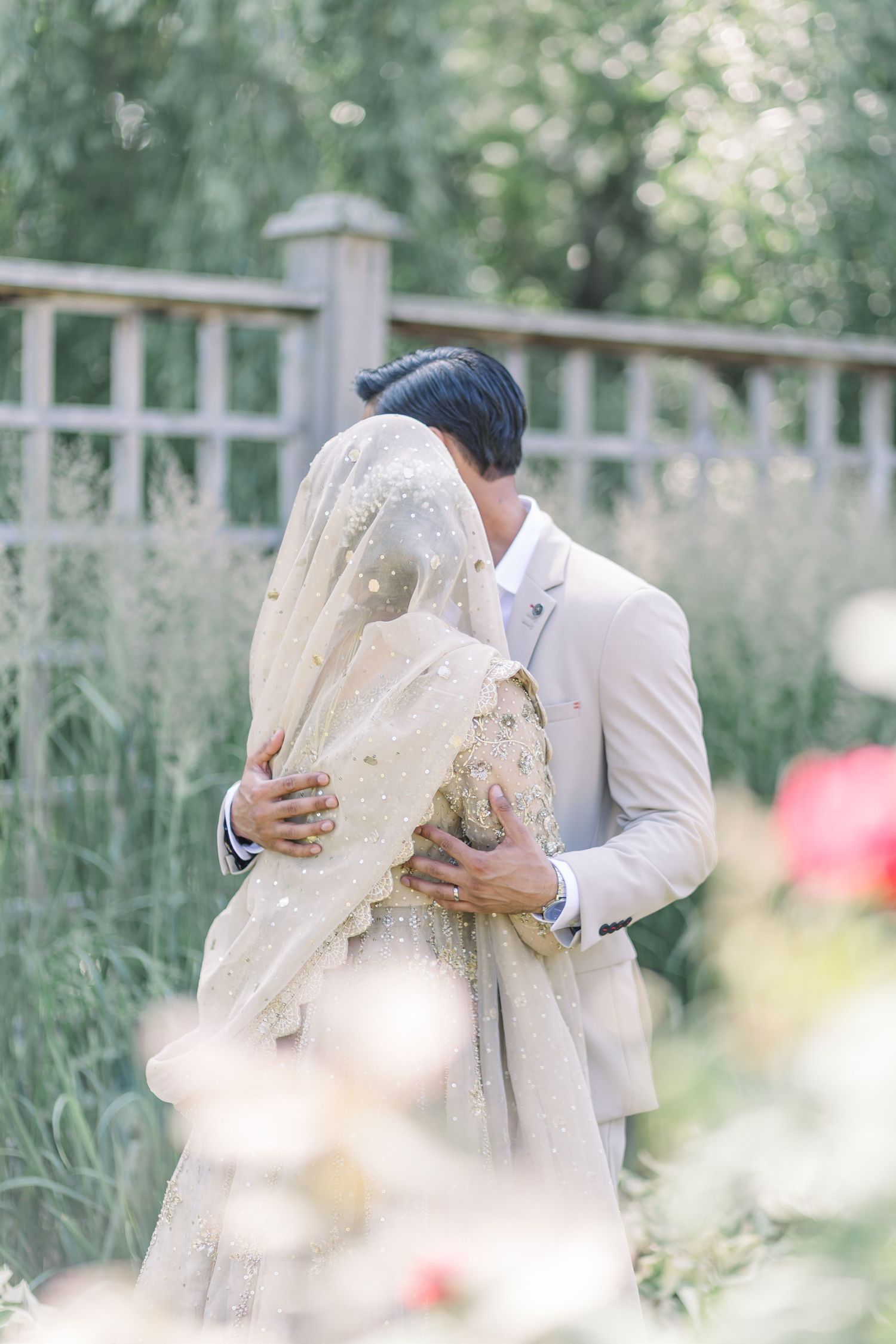 A romantic couple in white attire embraces in a dreamy garden setting with soft bokeh and flowers in the background.
