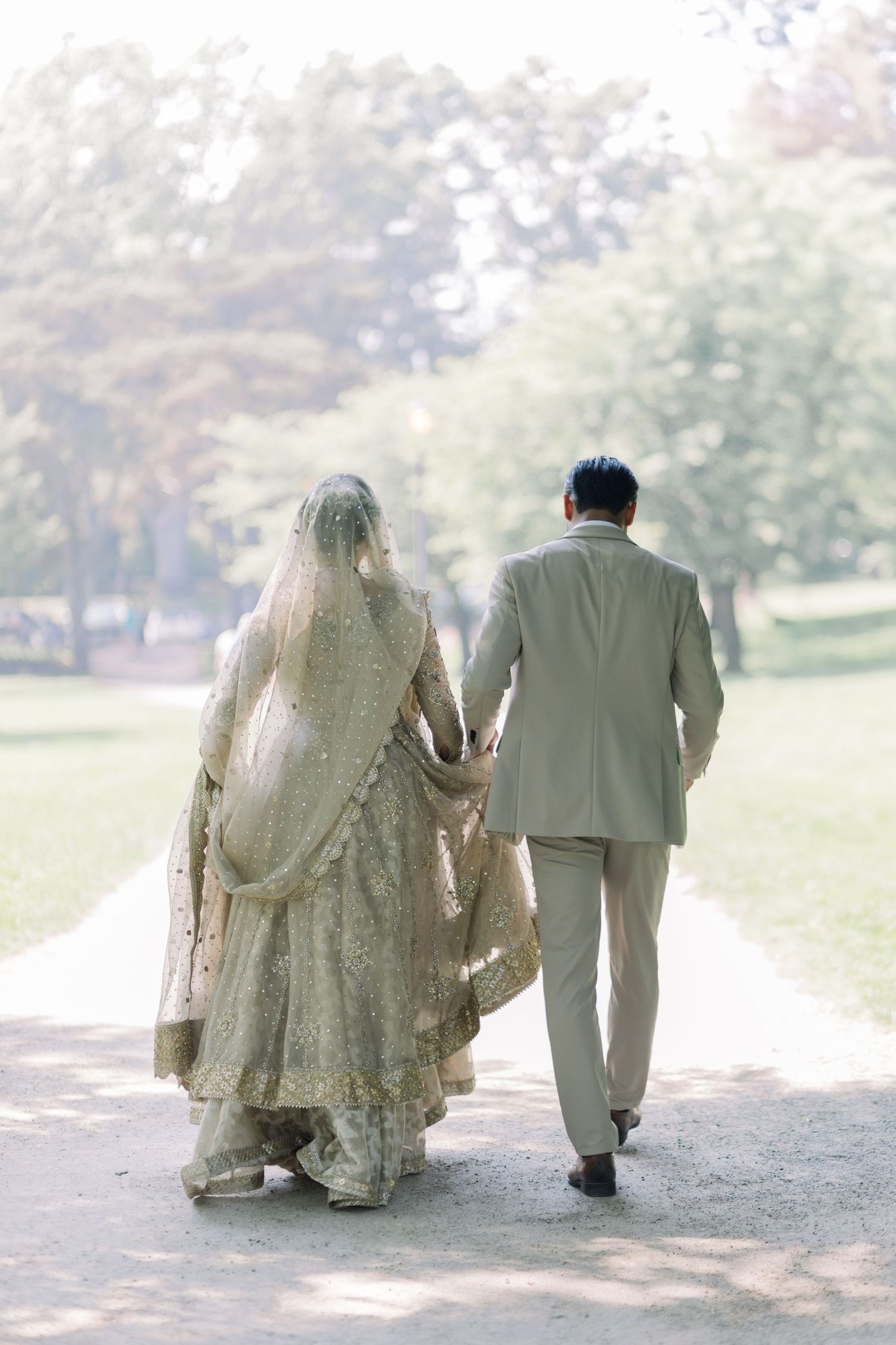 A couple in traditional wedding attire walks together along a scenic outdoor path in soft natural lighting.