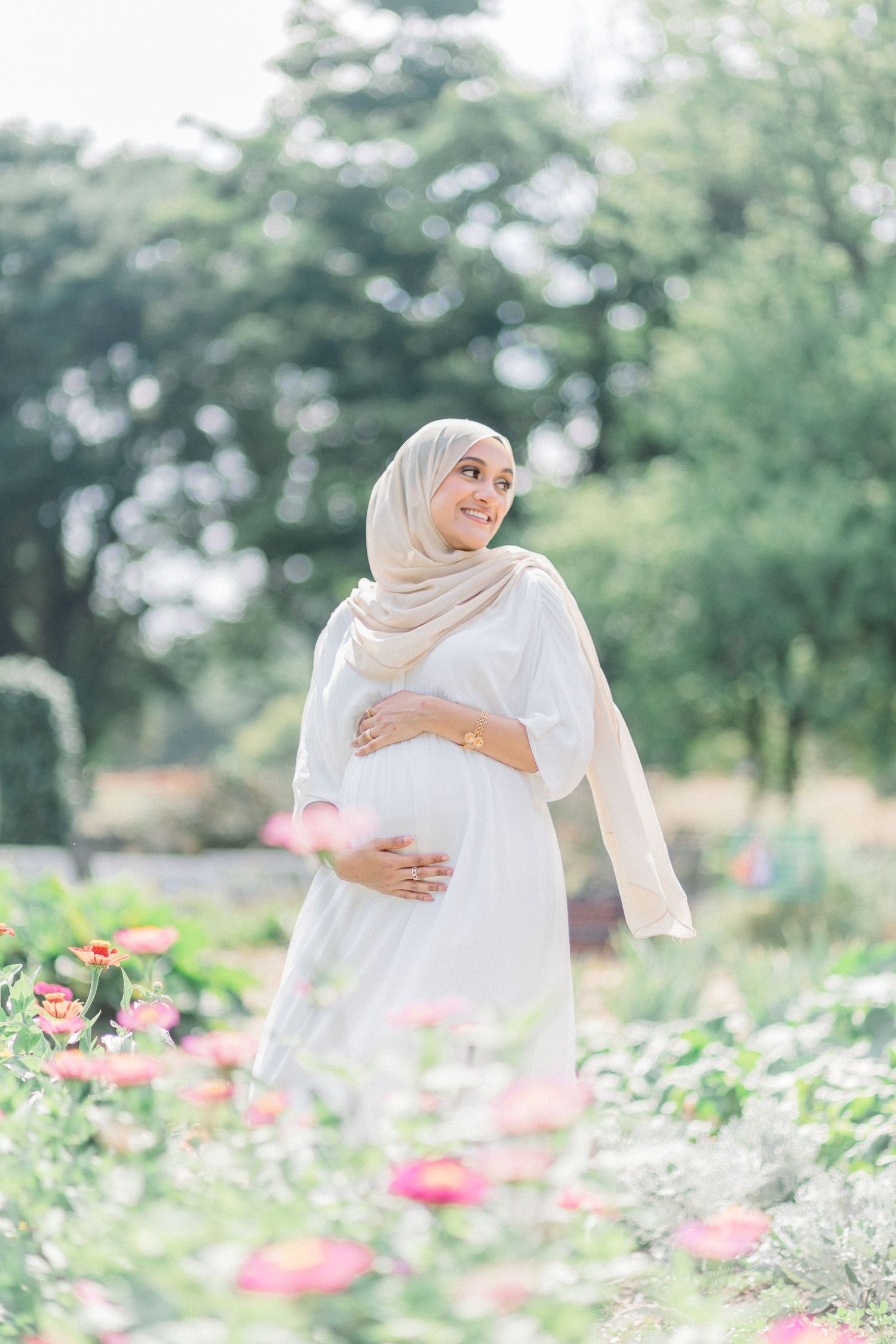 A person in a white hijab and dress poses gracefully in a blooming garden with pink flowers and greenery.