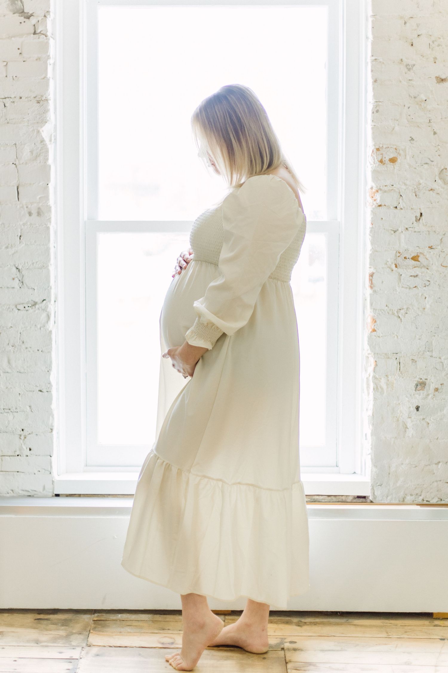 A person in a flowing cream-colored dress spins and twirls gracefully in a bright white room with exposed brick walls.