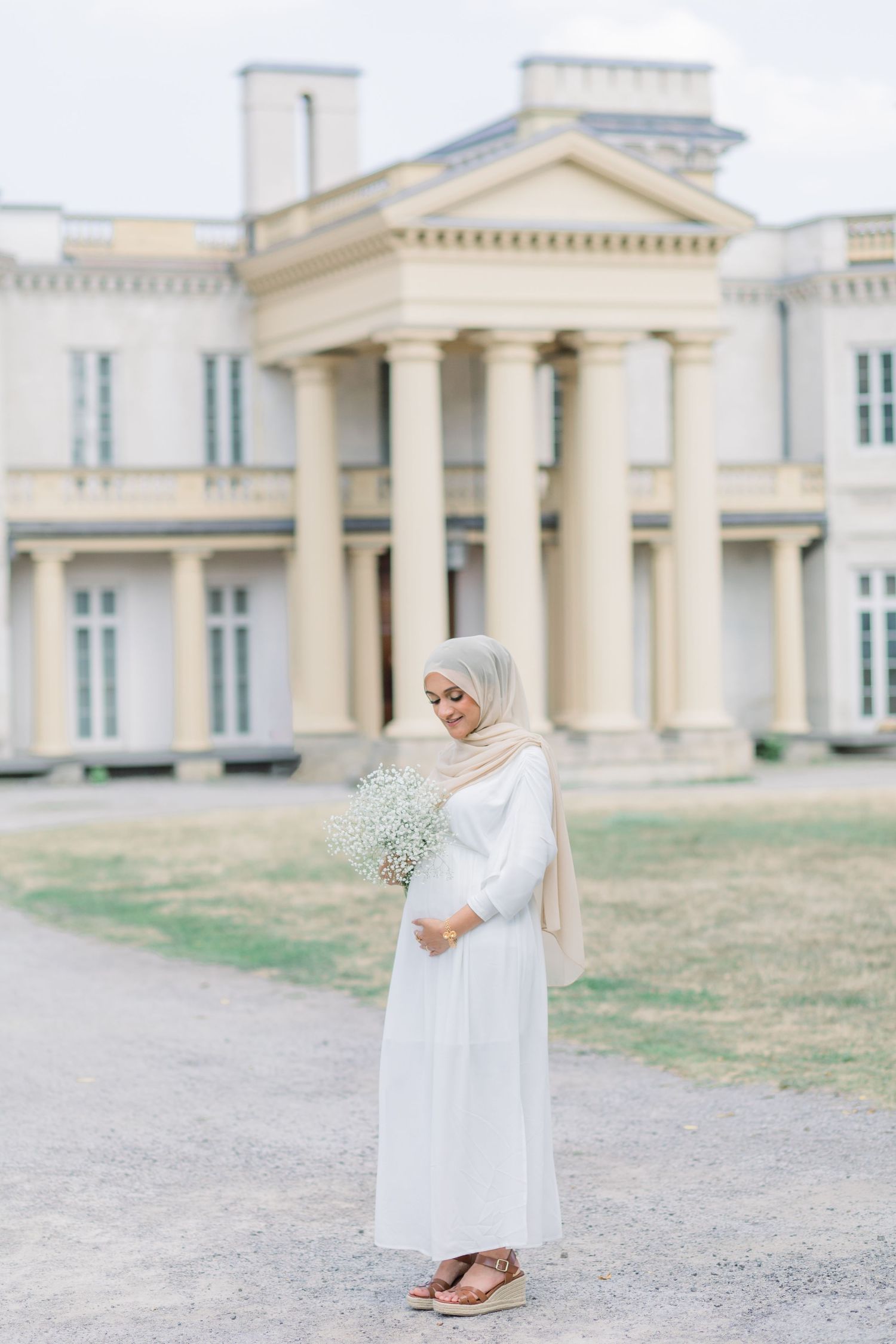 A person in flowing white dress stands gracefully against a neoclassical building with columns and pale walls.