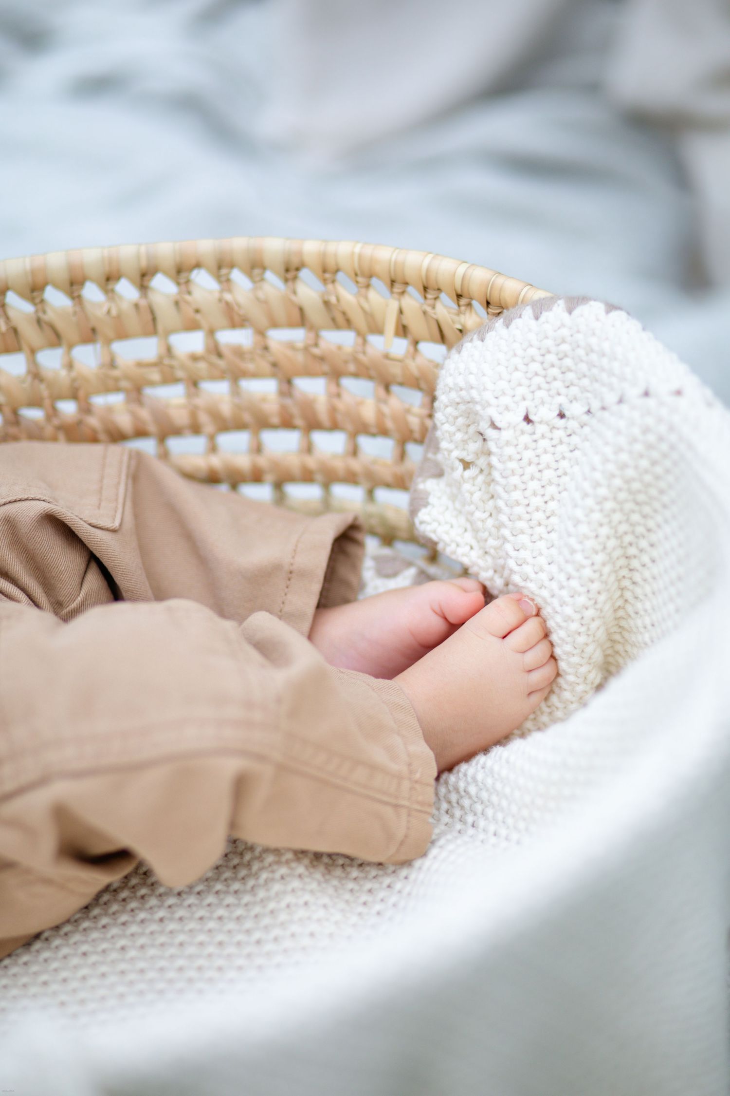 Close-up of tiny baby feet in tan pants resting on white knitted blanket in woven basket.
