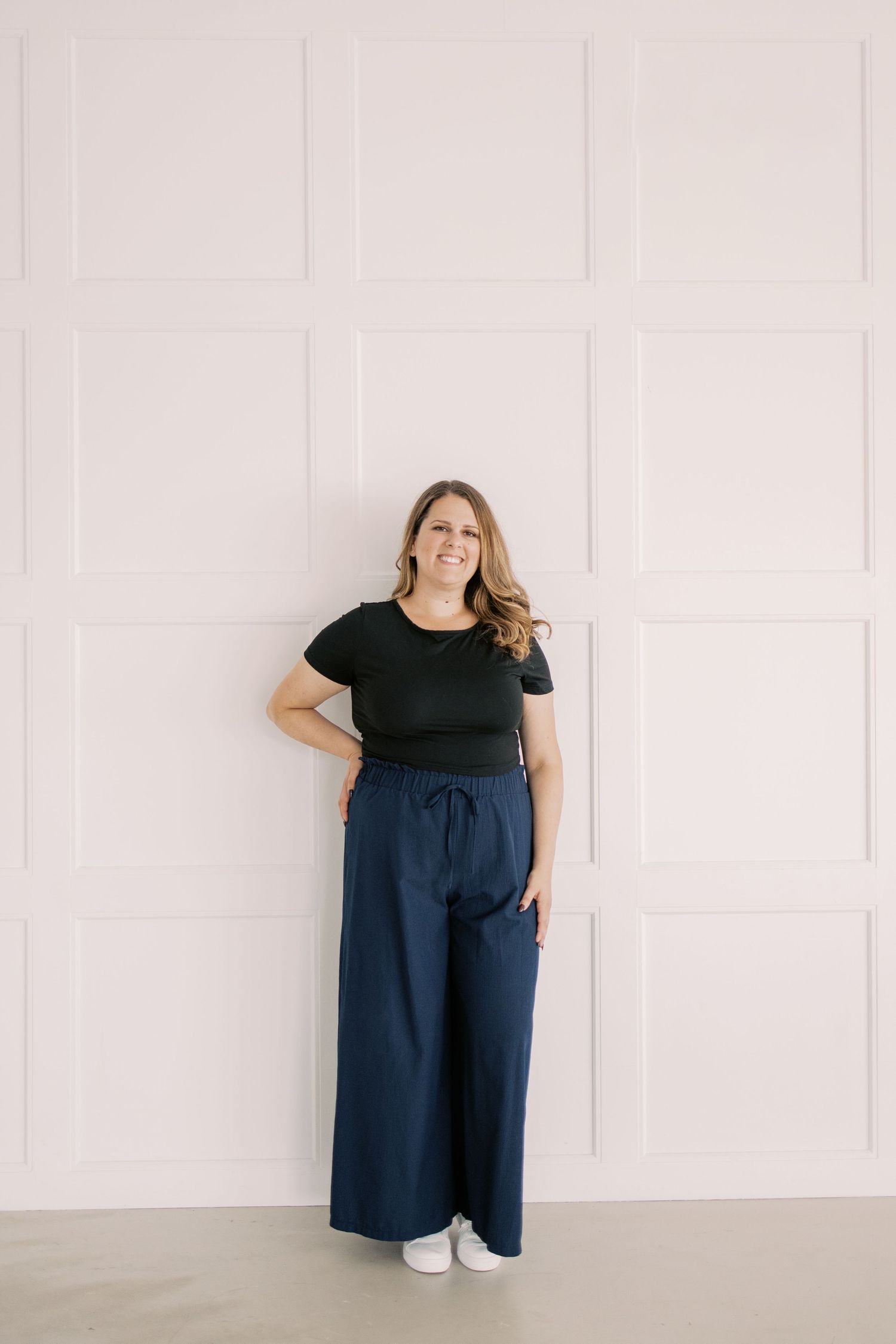 Woman wearing black t-shirt and navy pants poses against white wall in four casual lifestyle portraits.